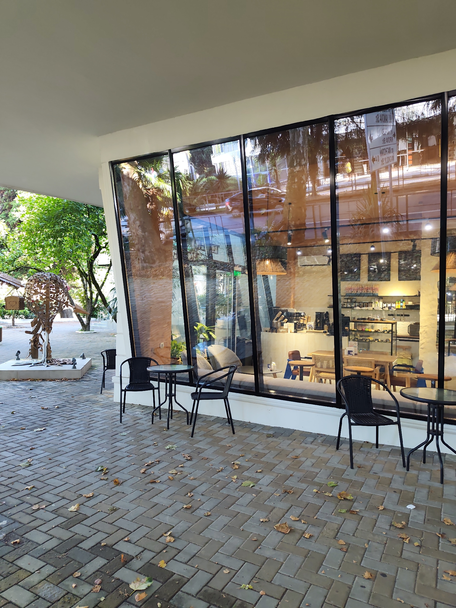 The image depicts the exterior of a cafe with large glass windows facing an outdoor seating area. The seating area is paved with gray interlocking bricks, and scattered autumn leaves are visible on the ground. Two small, round tables with black chairs are set up outside. Inside the cafe, several tables and chairs are visible, along with shelves stocked with merchandise and a coffee preparation area. Reflections in the glass show trees, a road, and cars, suggesting an urban environment. On the left side of the image, a large, abstract metal sculpture stands on a white base. The overall scene suggests a bright, possibly sunny day. This scene is located in Sochi, Russia.