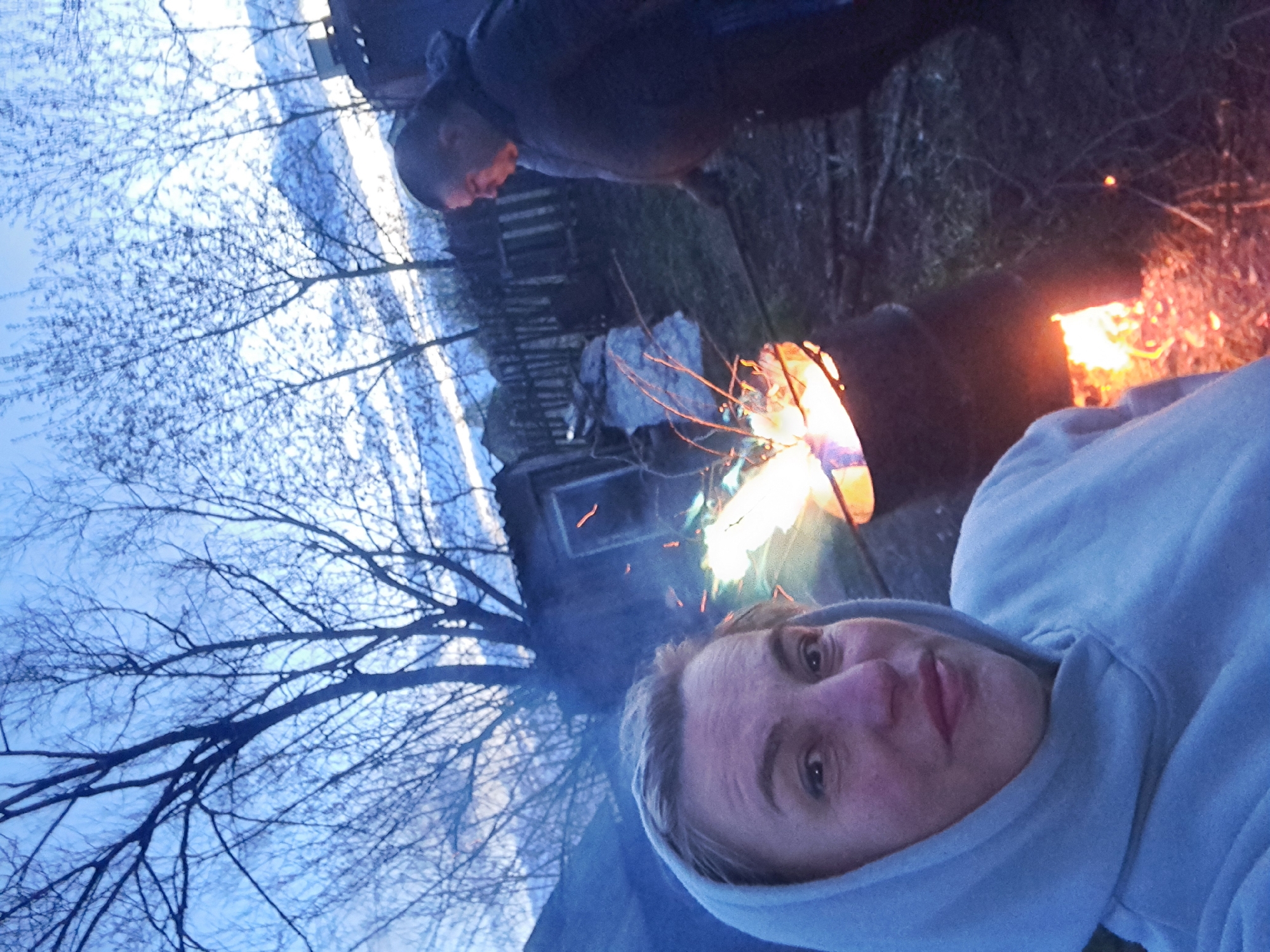 The image, originally rotated, depicts an outdoor evening scene in Полянки, Russia, featuring two individuals gathered around a crackling bonfire. In the immediate foreground, a person is taking a selfie, their face looking towards the camera with a neutral expression. They are wearing a light blue or grey hooded sweatshirt, with the hood up.
Behind and to the right of the foreground person, a bright bonfire illuminates the scene with vivid orange and yellow flames, and sparks are visible rising from it. Further back and slightly to the left of the fire, another person is visible in profile, wearing a dark jacket and looking down towards the fire.
The setting appears to be a rural or semi-rural area. Bare tree branches, characteristic of late winter or early spring, are silhouetted against a light blue, overcast sky that suggests dusk or early evening. In the mid-ground, behind the trees on the left, a rustic building or shed with a small window is partially visible. The ground is covered with what appears to be dry grass or earth. The overall atmosphere is one of a calm gathering around a warming fire as daylight fades. No visible text is present in the image.