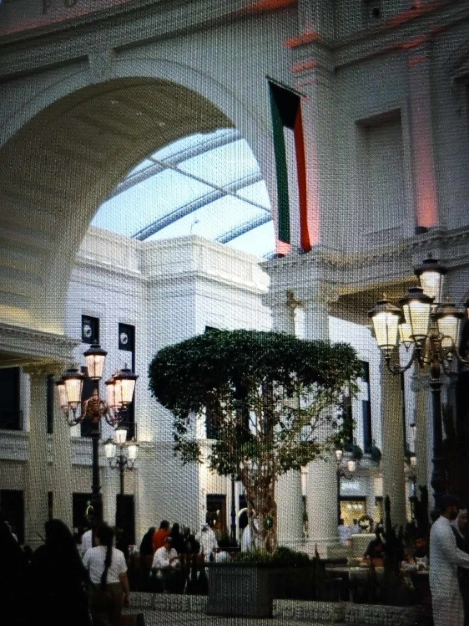 The image depicts an indoor atrium with a large arched opening covered by a glass ceiling. A Kuwaiti flag is displayed vertically on a white wall. In the foreground, a multi-tiered lamppost with illuminated lamps stands to the right of a potted tree. Several people are visible in the background, some gathered in groups. The architecture features white columns and intricate detailing. The overall scene suggests a public indoor space, possibly a shopping mall or a large governmental building. The reference to "Golbembal, Nigeria" appears to be an erroneous or irrelevant detail not supported by visual cues in the image.