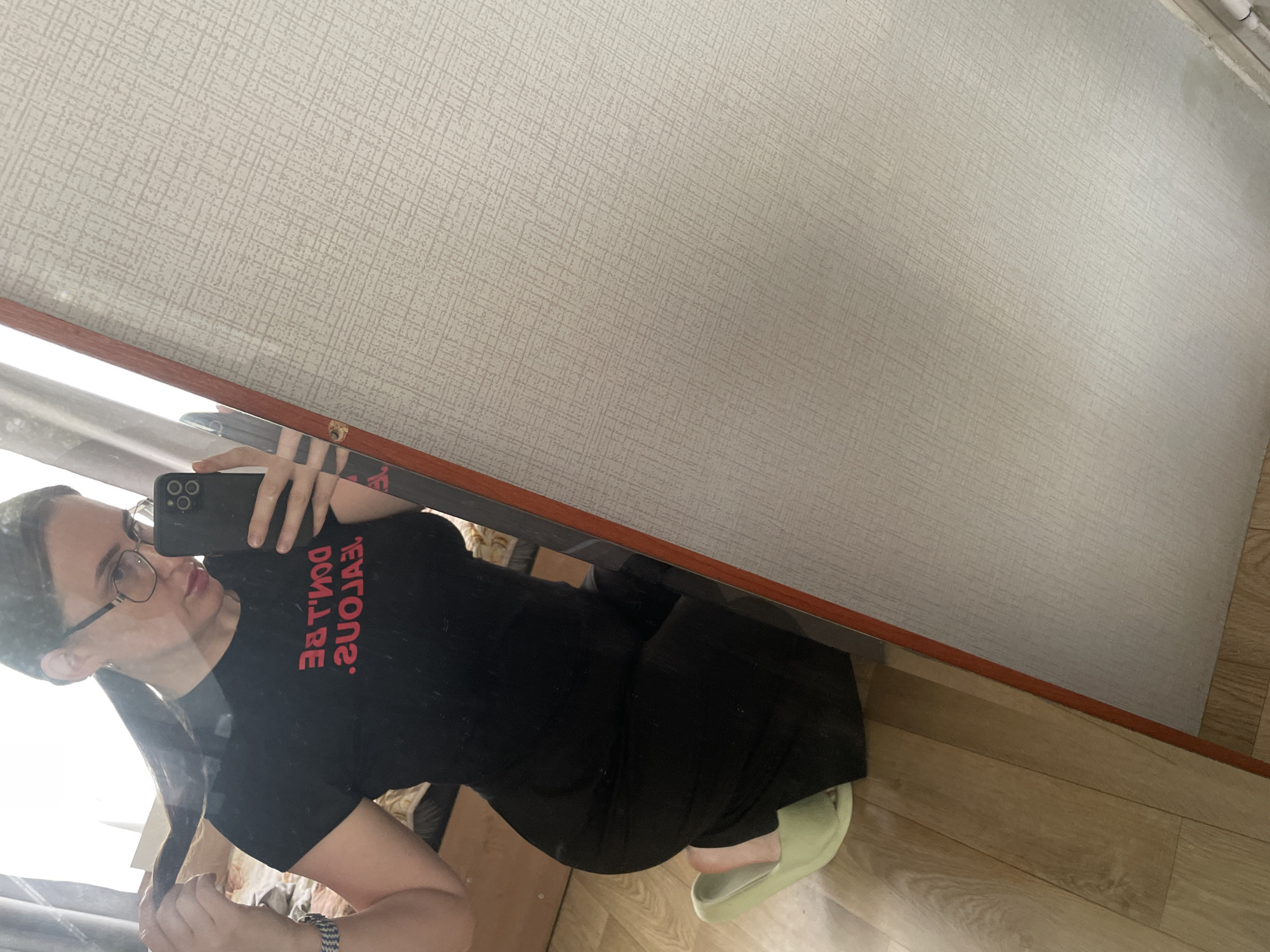 A woman wearing glasses and a black t-shirt with red lettering stands in a room. The lettering on her shirt reads "DON'T BE JEALOUS" in reverse. She is holding her hair in her left hand and a smartphone in her right hand, taking a selfie in a mirror. She is kneeling on a wooden floor, wearing light green slippers. The room appears to be a bedroom, with a bed visible in the mirror's reflection, and wallpaper on the wall. The location is noted as being in the city of Сырково, Russia.