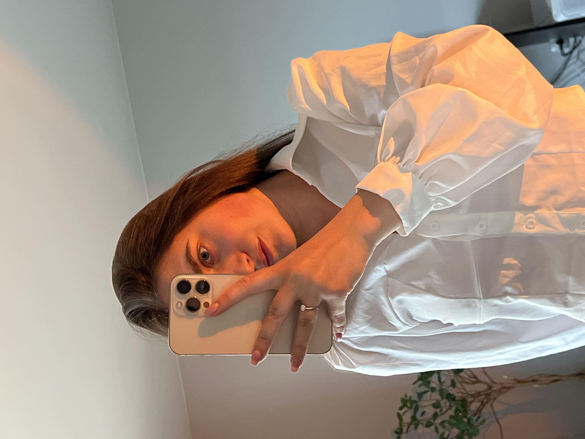 The image displays a mirror selfie taken by a woman in an indoor setting in Ternopil, Ukraine. The woman, with dark brown hair, is captured from the chest up, with her head slightly tilted. Her right eye, nose, and lips are visible as she looks directly at the camera with a neutral and attentive expression.
She is wearing a white collared shirt featuring prominent, voluminous puffy sleeves that gather at the cuffs. The fabric of the shirt shows soft folds and creases, indicating a relaxed fit. Her right hand is extended, holding a light-colored smartphone with a distinctive triple-camera array, suggesting an iPhone. A ring with a small, light-colored stone is visible on her right ring finger, and her nails are painted with a subtle, light polish.
The background consists mainly of light-colored walls, possibly off-white or light grey, which reflect a warm, golden light. This illumination bathes her skin, arm, and the white shirt, creating a soft, inviting glow. In the lower right corner of the frame, a houseplant with green leaves and slender bare branches is partially visible, adding a touch of natural greenery to the scene. The overall lighting suggests either morning or late afternoon. No visible text is present in the image.
