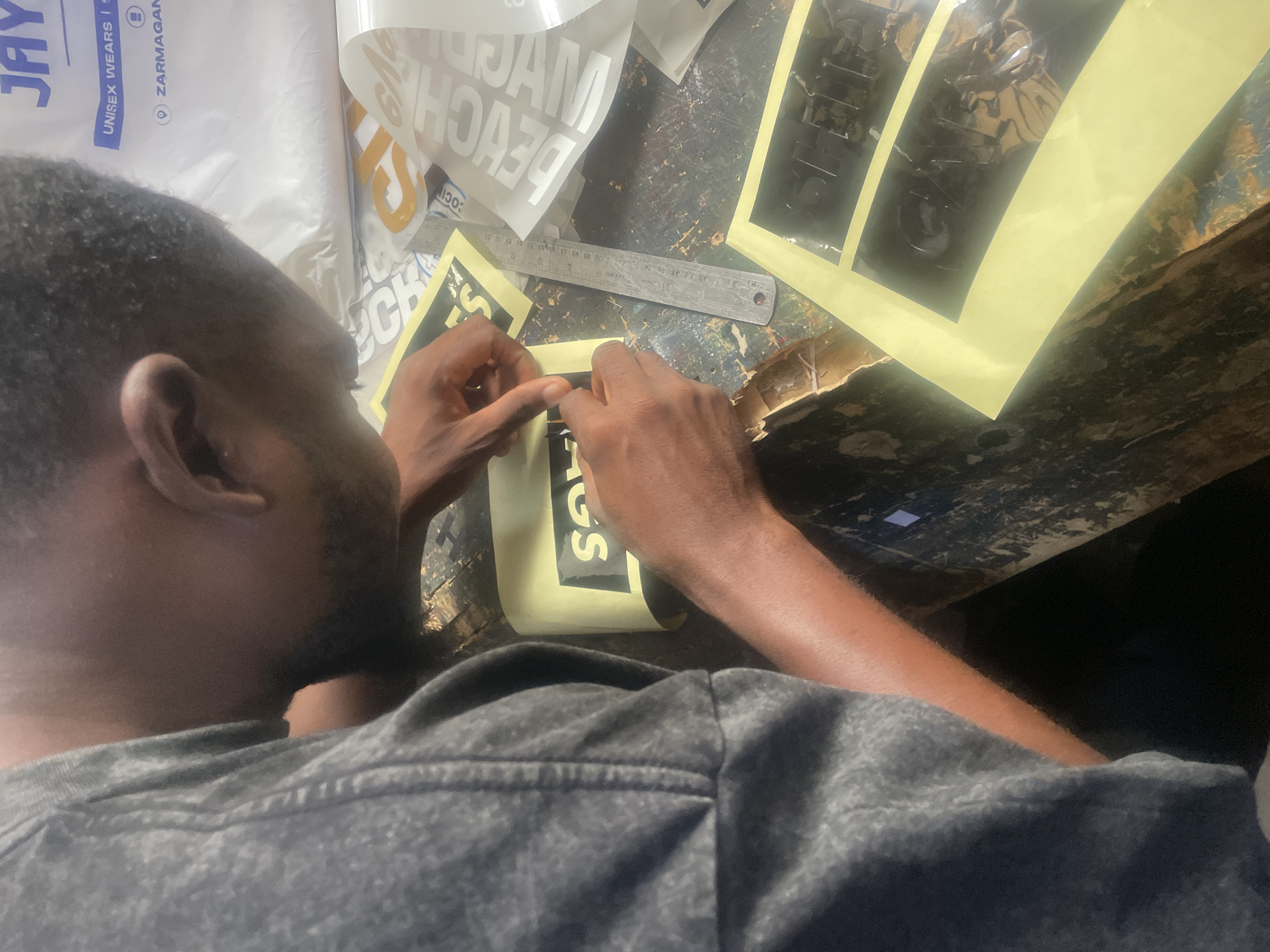 The image shows a close-up of a person's hands as they work on a craft project, likely in a workshop setting. The individual, whose face is only partially visible, is wearing a gray t-shirt. Their hands are carefully manipulating a yellow sticker with black lettering. Adjacent to this sticker are several other decals, some with white lettering on a clear background and others with black lettering on a transparent film. A metal ruler is placed on the work surface, suggesting precision is involved in the task. The background appears to be a rustic, possibly wooden table with a textured, speckled surface, indicating a workshop or craft area. The overall lighting suggests an indoor environment. Visible text on various items includes "JAY," "UNISEX WEARS IS," "ZARMAGAN," and what appears to be "BENCH" and "SOC IN." The context provided indicates the location is Jos, Nigeria. The activity seems to be the application or preparation of vinyl decals.
