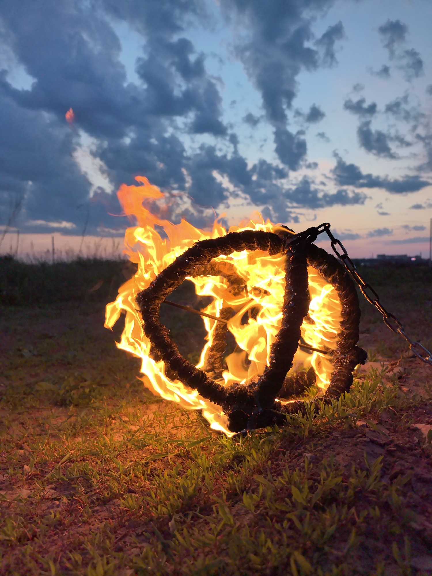 A ground-level, eye-level shot captures a spherical fire performer's prop engulfed in flames against a twilight sky with cloud cover. The prop is composed of two intertwined rope circles connected by thin metal rods, with a short metal chain extending from one of the circles. The prop is set on a patch of green grass and reddish-brown soil. In the background, distant trees and buildings are visible along the horizon. Based on the prompt, the location is in or near Фролы, Russia.