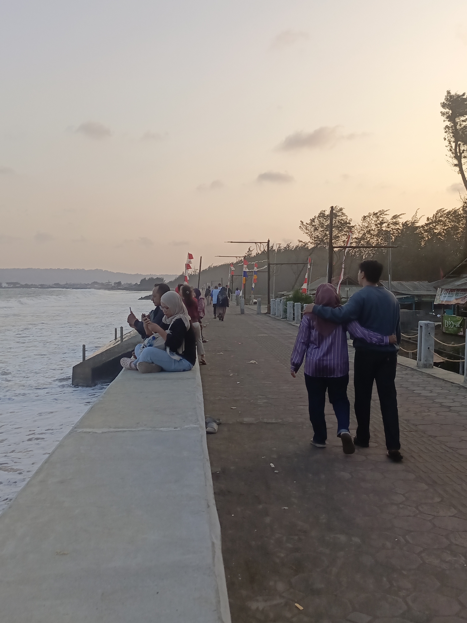 A couple walks arm-in-arm along a paved walkway next to the ocean in Kelurahan Tegalkamulyan, Cilacap, Indonesia. The walkway runs alongside a concrete sea wall, with foamy waves visible on the left. The sky is overcast with a pale yellow and pink hue, suggesting either dawn or dusk. Several other people are present, some seated on the sea wall and others walking further down the path. Small structures with flags are visible on the right side of the walkway, indicating commercial activity or local decorations. Trees line the background behind the structures. The overall atmosphere is casual and relaxed, with people enjoying the coastal scenery.