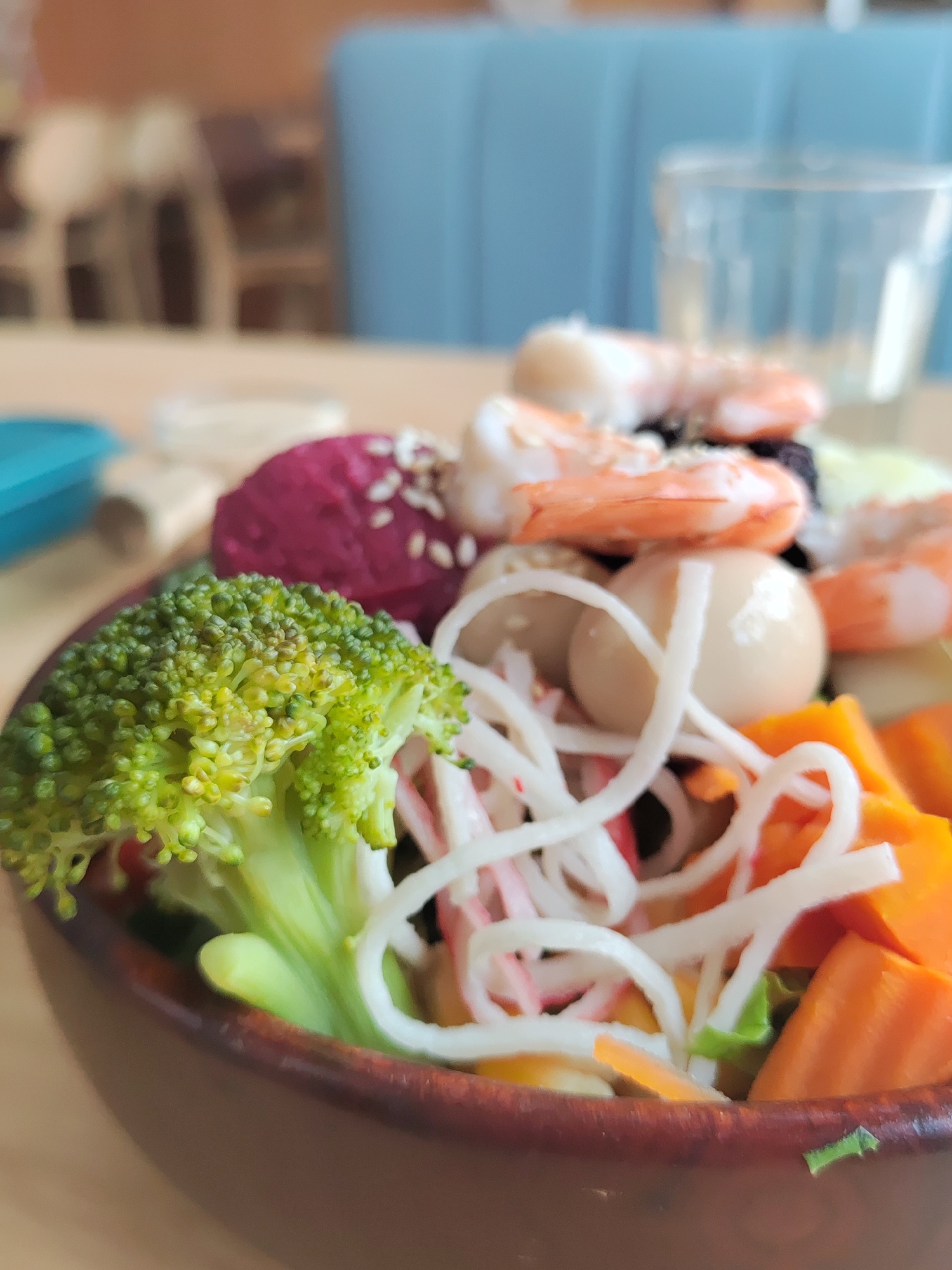 A close-up shot shows a bowl of food, likely a salad, filled with various ingredients. Prominently visible are broccoli florets, shredded imitation crab meat, diced carrots, boiled quail eggs, a beetroot ball with sesame seeds, and cooked shrimp. The bowl appears to be made of dark brown ceramic. In the blurred background are a light-colored wooden table, a light blue upholstered chair, and a glass of liquid. The photo was likely taken indoors. While it is possible this could be Da Lat, Vietnam, no specific elements within the frame conclusively confirm this location.