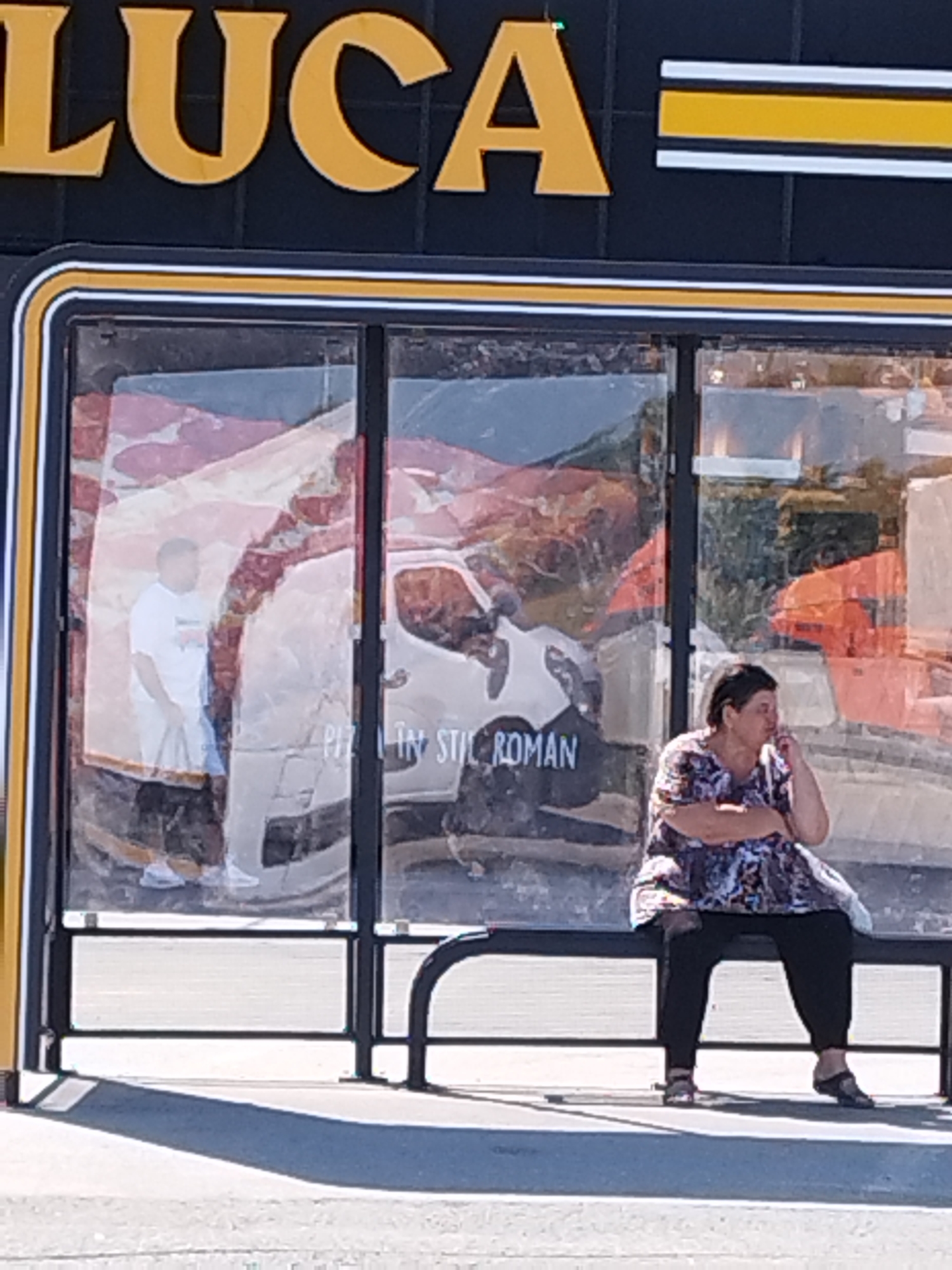 An outdoor shot captures a bus stop in Bragadiru, Romania. A woman in a floral-patterned top and black pants sits on a bench, her hand near her face, appearing to be holding a cigarette. Behind her, through the glass panels of the bus stop, a large advertisement for pizza is visible, featuring images of pizza and a partially obscured picture of a man in a white t-shirt and blue shorts. The advertisement also contains text that reads "PIZZA IN STIL ROMAN". The bus stop structure itself is framed by black metal poles and a bench. The setting appears to be a sunny day, with shadows cast on the pavement. Above the bus stop, a sign with large yellow letters and black and white stripes is visible, with the letters "LUCA" clearly legible.