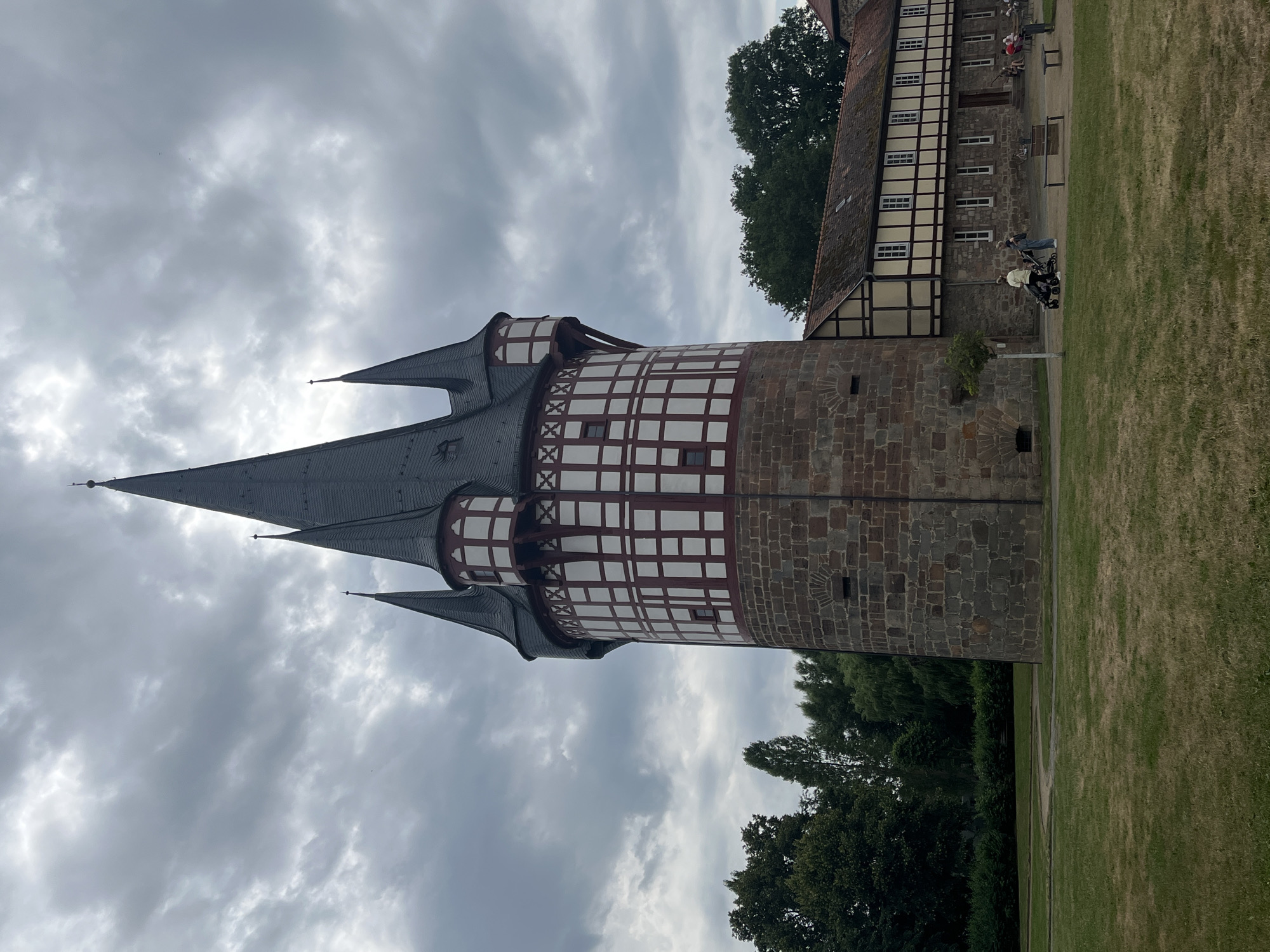 This image captures a view of a historical tower with a distinctive half-timbered upper section and a stone base. The tower features multiple conical roofs, the tallest of which is a dark gray slate. The half-timbered section is white with dark red beams, forming a crisscross pattern.
To the right of the tower, a building with similar half-timbered architecture is visible. This building has a red-tiled roof and multiple windows. The area around the tower and building is a grassy lawn.
In front of the building, several people are seen. Two individuals are pushing strollers, and a few others are seated on a bench or in the background. The scene suggests a daytime visit to a historical site or park. The sky is overcast with gray clouds, indicating cloudy weather.
The location is Neustadt, Germany, which is suggested by the architectural style commonly found in this region. The overall atmosphere is peaceful and leisurely, with visitors enjoying the grounds. There is no visible text in the image.
