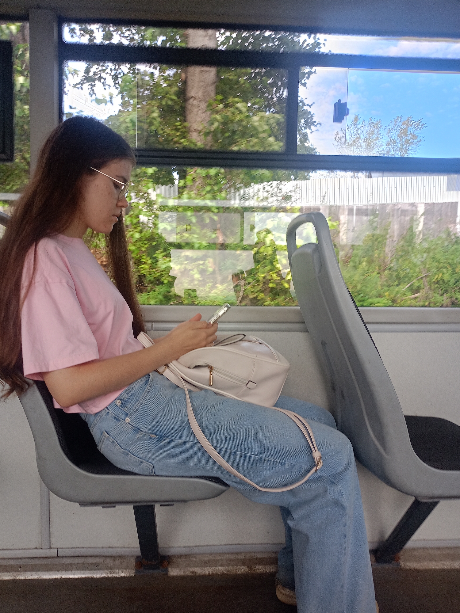 A young person with long brown hair and glasses, wearing a pink t-shirt and blue jeans, is sitting on a gray bus seat, looking at their phone. A light-colored purse is resting on their lap. The bus is moving, as indicated by the blurred greenery and blue sky visible through the windows. The scene takes place inside a public transport vehicle, likely a bus, which appears to be traveling on a route in or near Zlatoust, Russia, given the surrounding vegetation and the architectural style of the fencing visible outside.