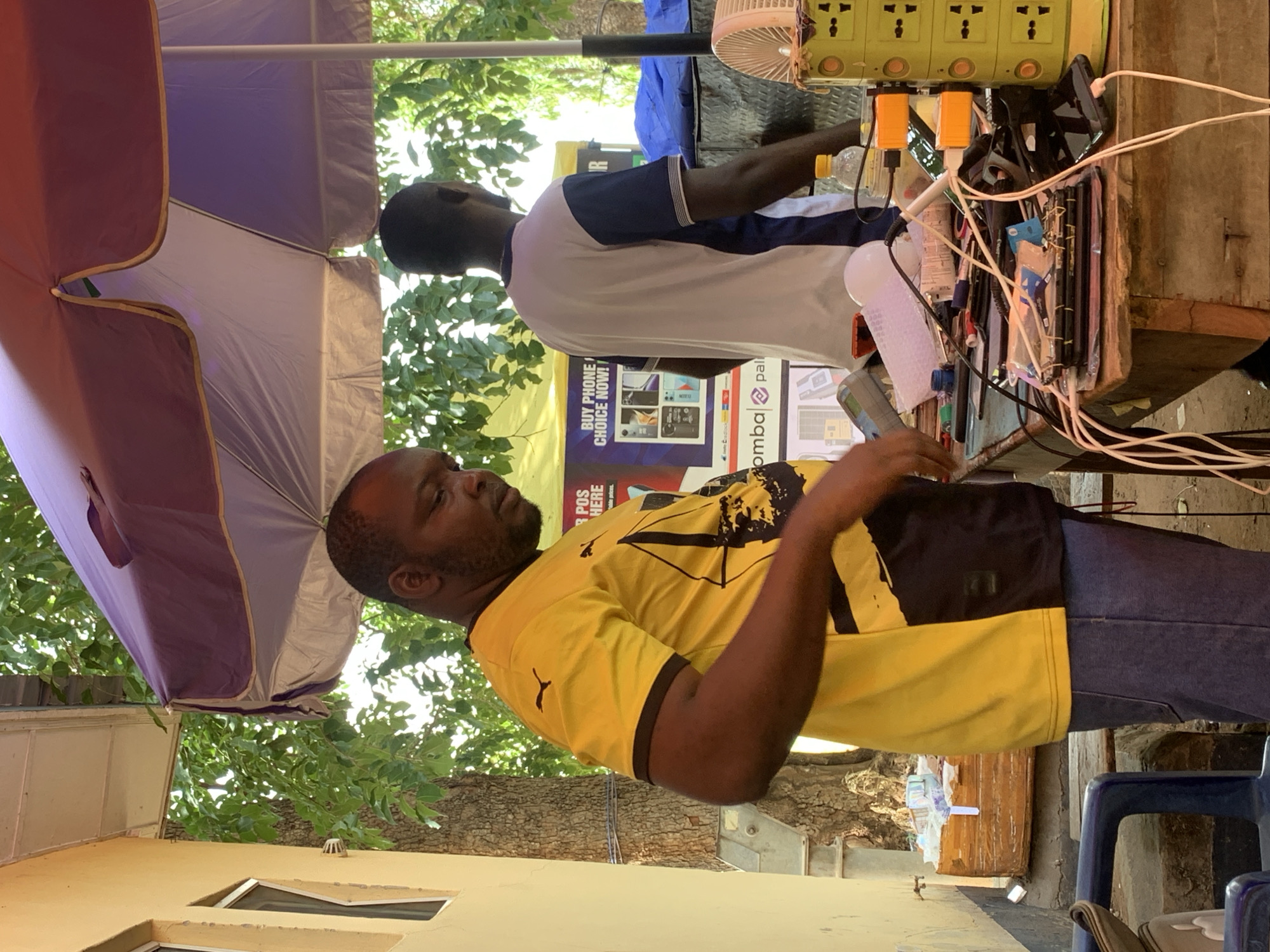 This media file captures an outdoor or semi-outdoor scene, likely a public phone charging station in Ilorin, Nigeria, under bright daylight.
In the foreground, a man with a beard, wearing a yellow and black t-shirt (with a visible Puma logo) and dark blue jeans, stands looking towards the left. He is holding a small, gray handheld device, possibly a meter or scanner. Behind him, another man, seen from the back, wears a white polo shirt with navy blue sleeves and collar. He is attending to a collection of electronic devices and cables arranged on a wooden surface.
This setup includes a green multi-socket power strip with several yellow adapters plugged in, feeding numerous white charging cables that connect to multiple smartphones and other gadgets. A small pink fan and a white lightbulb are also visible on the table. The presence of these items strongly suggests a service offered for charging mobile phones and electronic devices.
An open purple and white umbrella provides shade over the charging station, indicating sunny weather. In the background, lush green trees and foliage are visible, along with a light-colored building wall. A blue plastic chair is partially seen in the lower right.
A yellow banner or sign is displayed behind the charging station, featuring images of different smartphones. Visible text fragments on the banner include "BUY PHONE...", "CHOICE NOW!", "POS HERE", and "omba | pal". The scene depicts a typical small-scale service business catering to public needs in a local setting.