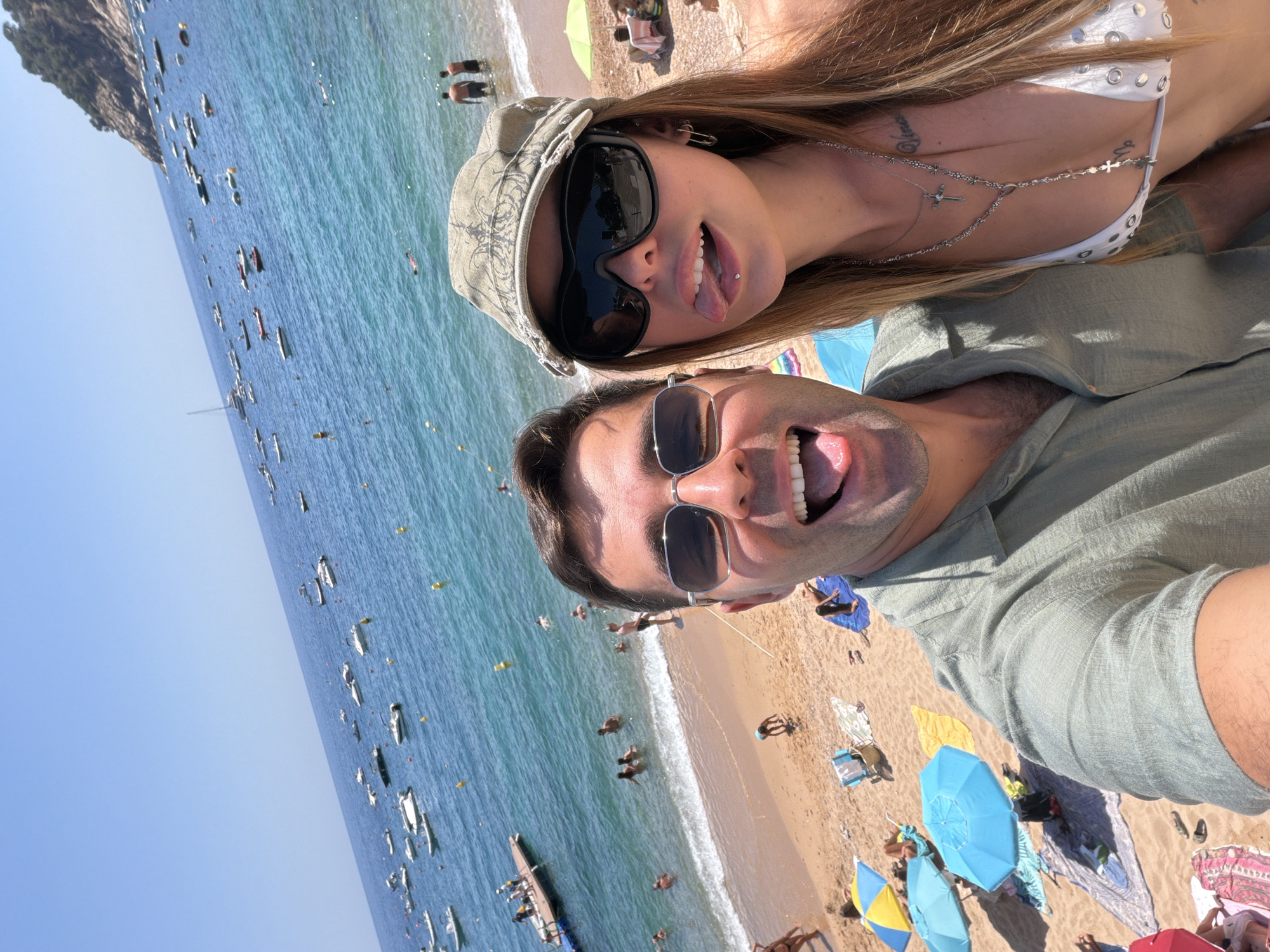 This is a close-up, selfie-style photograph taken on a sunny day at the beach in Tossa de Mar, Spain. The foreground features a man on the left and a woman on the right, both looking towards the camera with open mouths. The man is wearing a light green short-sleeved shirt and sunglasses. The woman is wearing a white bikini top, large dark sunglasses, and a cap. She is sticking her tongue out and has a lip piercing. Behind them, the sandy beach is populated with several beach umbrellas and people sunbathing or swimming in the turquoise water. Numerous boats are visible anchored offshore under a clear blue sky.