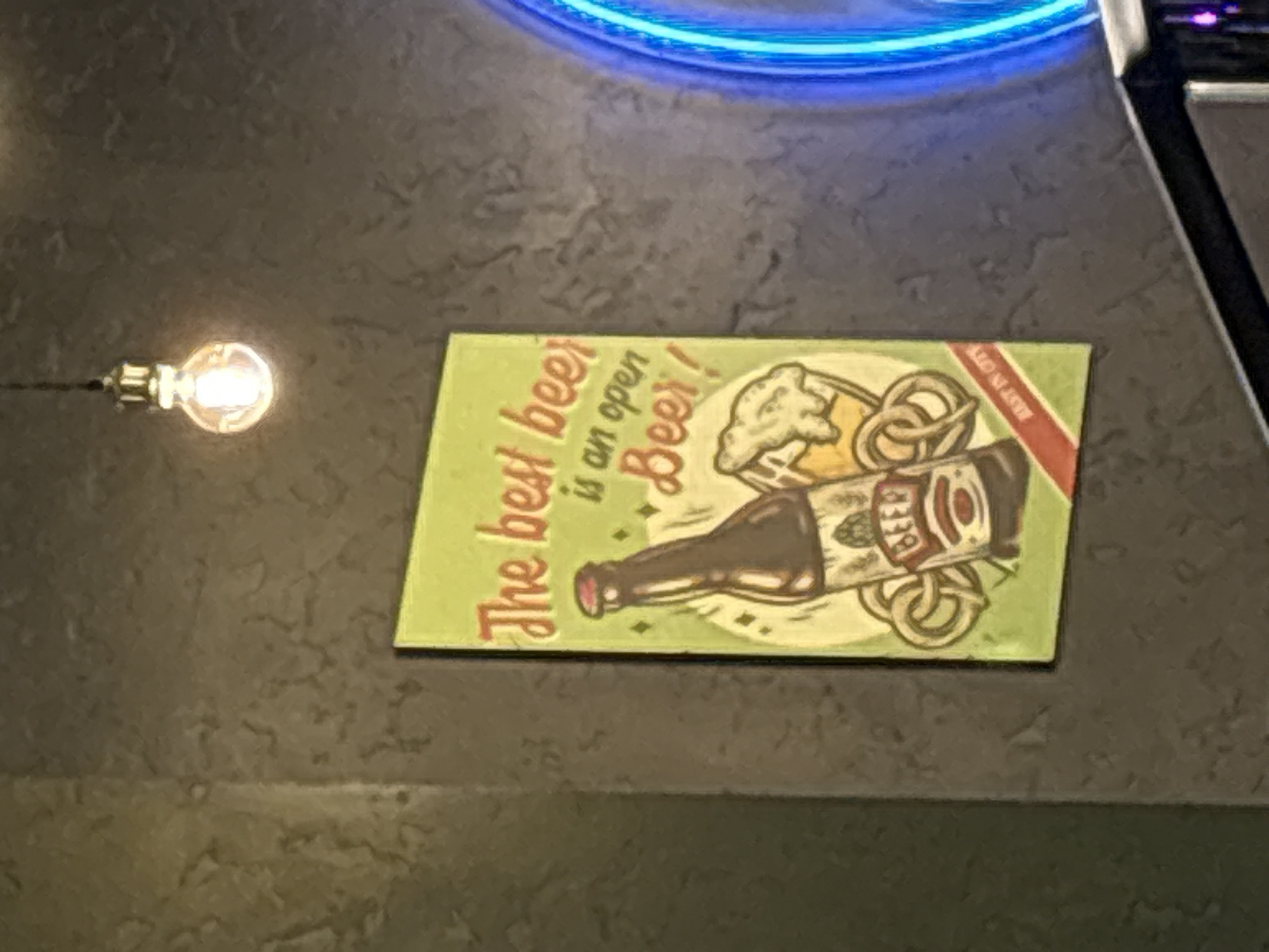 The image displays an indoor scene, likely within a bar or restaurant in Izhevsk, Russia, characterized by a dark, textured surface, possibly a bar top or table. A rectangular sign or coaster, designed in a retro or vintage style, is positioned centrally on this surface.
The sign features an illustration of a brown beer bottle lying horizontally, accompanied by a frothy mug of beer and two pretzels. The dominant colors of the sign are light green, yellow, and muted browns. Prominently displayed text on the sign, written in a cursive font, reads: "The best beer is an open Beer!". A smaller, partially obscured red banner on the right side of the sign contains some text, though it is difficult to decipher fully. The word "BEER" is also visible on the label of the illustrated bottle.
To the left of the sign, a single, glowing light bulb hangs by a wire, emitting a warm, bright light that illuminates the immediate area. Above the sign and slightly to the right, a vibrant blue neon light is partially visible, adding a cool-toned glow to the scene. On the far right edge of the frame, a dark, metallic object, possibly part of a fixture or piece of equipment, is partially captured. The overall dim lighting, coupled with the neon and bulb lights, suggests an evening or nighttime setting. No people are present in the image.