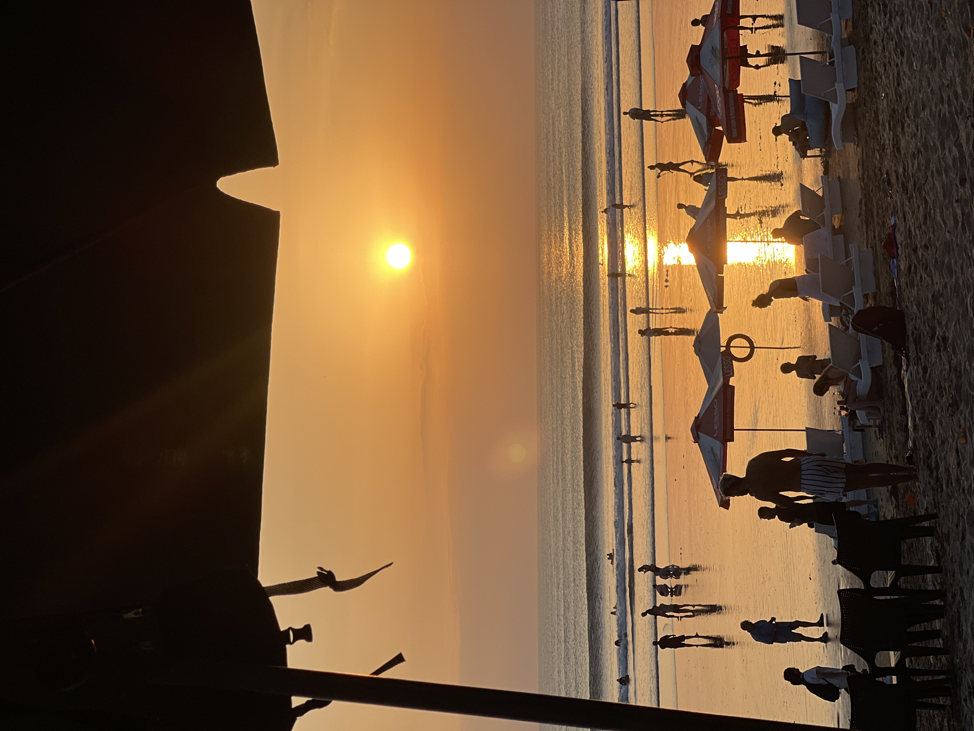 A group of people are enjoying a sunset at Kuta Beach in Kuta, Indonesia. The sun is low on the horizon, casting a warm, golden glow over the scene. The ocean is calm, with gentle waves lapping at the shore. Several people are walking along the beach, while others are wading in the water. Beach umbrellas and chairs are set up, providing shade for sunbathers. Silhouettes of people are visible against the bright sky. The overall mood is peaceful and relaxed.