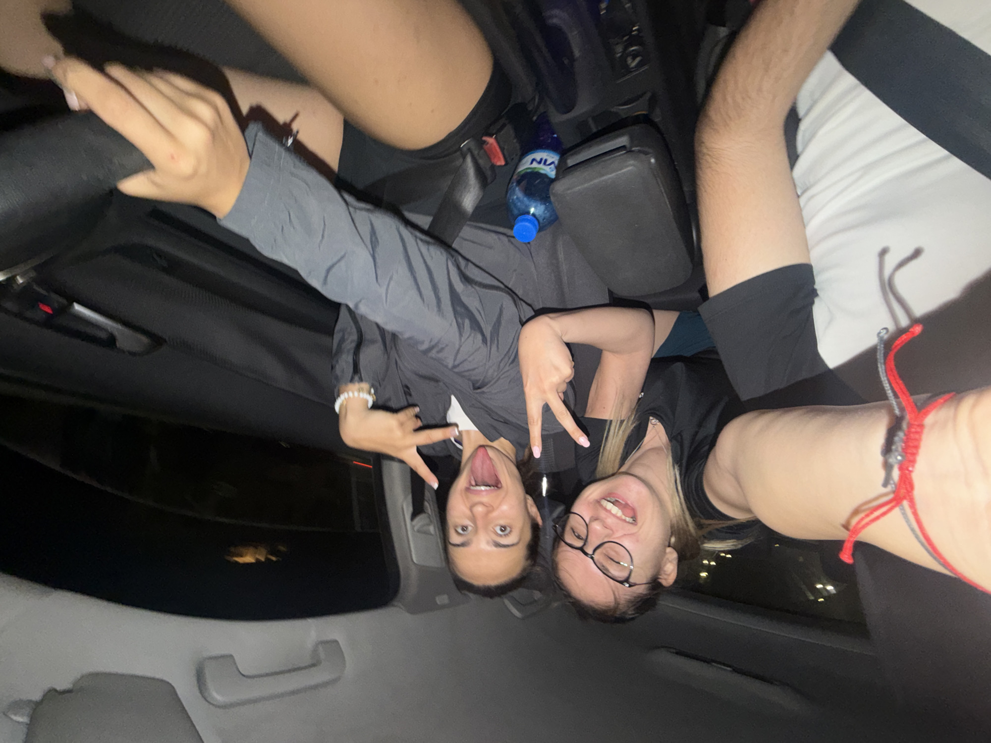 The image depicts three individuals inside a vehicle during nighttime. A female driver is positioned on the right, her left hand on the steering wheel and her right hand displaying a "V" sign. She wears a grey jacket over a white top and black shorts, with a white beaded bracelet on her left wrist. Her face shows wide-open eyes and an extended tongue. To her left, a female front passenger, wearing glasses and a black t-shirt, is captured mid-laugh with her head tilted. Her right arm is bent at the elbow, positioned near the central console. A third individual, likely the photographer, is partially visible in the left foreground, showing a right arm wearing a red and grey braided string bracelet, and a torso clad in a white t-shirt with black sleeves, secured by a black seatbelt.
The vehicle's interior is dark, featuring a black steering wheel, a dark grey central armrest, and visible seatbelt buckles. A blue plastic bottle labeled "VIN" with a blue cap is situated in a cup holder on the central console. A grey overhead grab handle is visible above the driver's side rear passenger window area. The exterior background through the windows is dark, showing blurred yellow lights in the distance and a faint red light visible through the driver's side window. The presence of a "VIN" branded water bottle, a product commonly found in Bulgaria, indicates the location. Based on this evidence and the prompt, the scene is situated in the city of Sofia, Bulgaria.