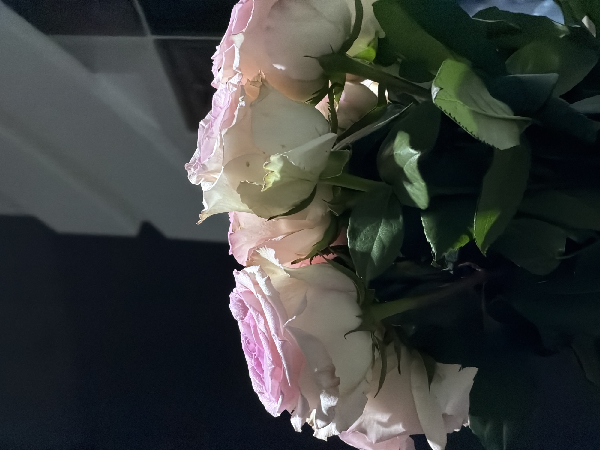 The image shows a close-up of a bouquet of light pink and white roses with green leaves. The lighting is dim, casting shadows on the background, which appears to be an indoor setting with dark walls and reflective surfaces. No specific details indicate the location within Khmelnytskyi, Ukraine.