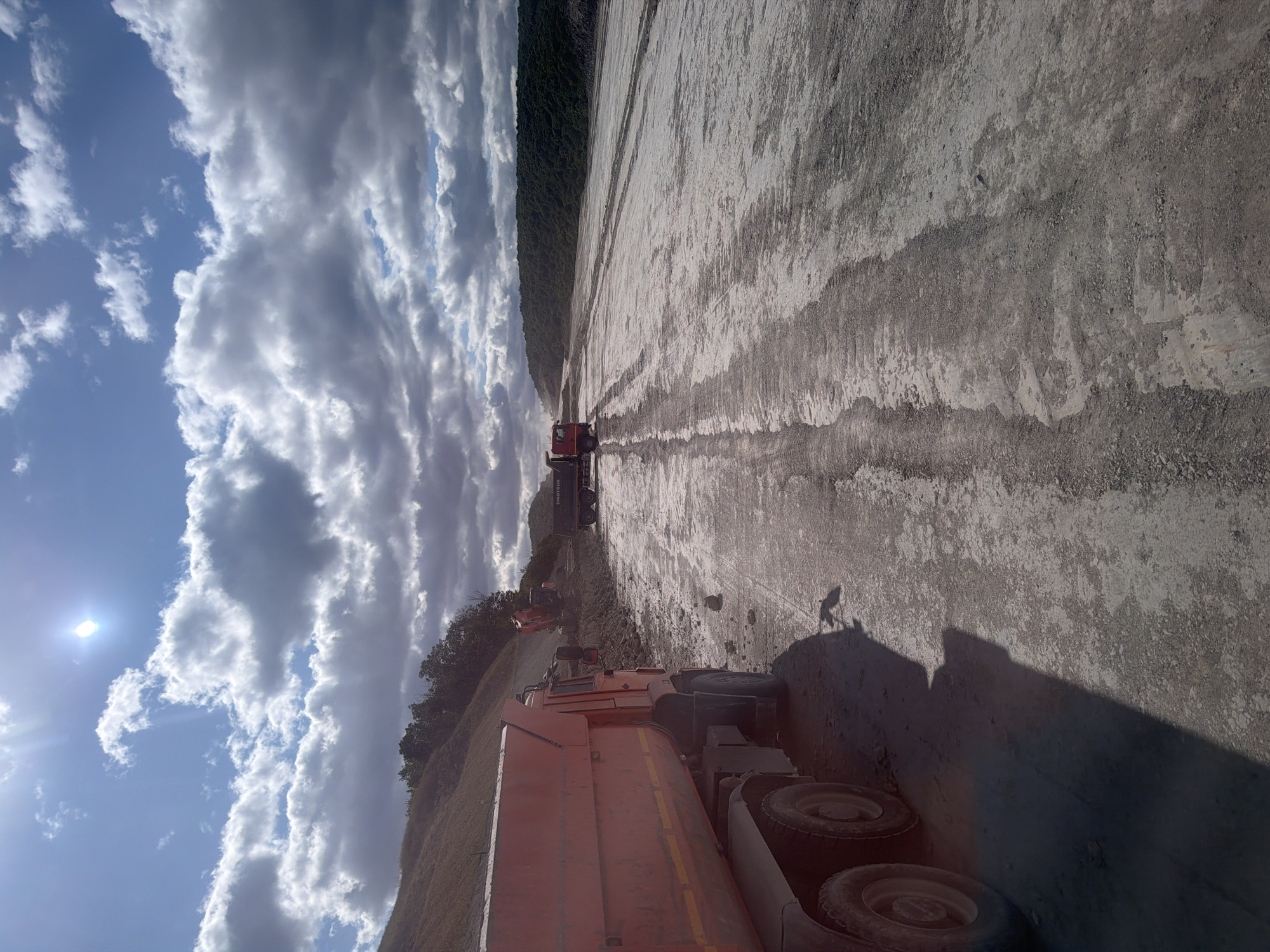The image depicts an outdoor construction or excavation site under a bright, partly cloudy sky. In the foreground, a large orange dump truck is parked on a dirt road. Its rear section is elevated, suggesting it might have recently dumped its load or is preparing to do so. The truck's wheels and undercarriage are visible. Further into the scene, on the right side of the road, is a long, cleared area with visible tire tracks, indicating heavy vehicle traffic. In the middle ground, another dump truck, this one dark-colored with "SINOTRUK" branding, is visible at a distance, positioned near an excavation area where an orange excavator is actively moving dirt. The background shows a sloping hill on the left, covered with sparse vegetation, and a dense line of trees forming a treeline on the right horizon. The overall environment appears to be a large-scale earthmoving operation. The location is identified as Kolotov Buryak, Russia.
