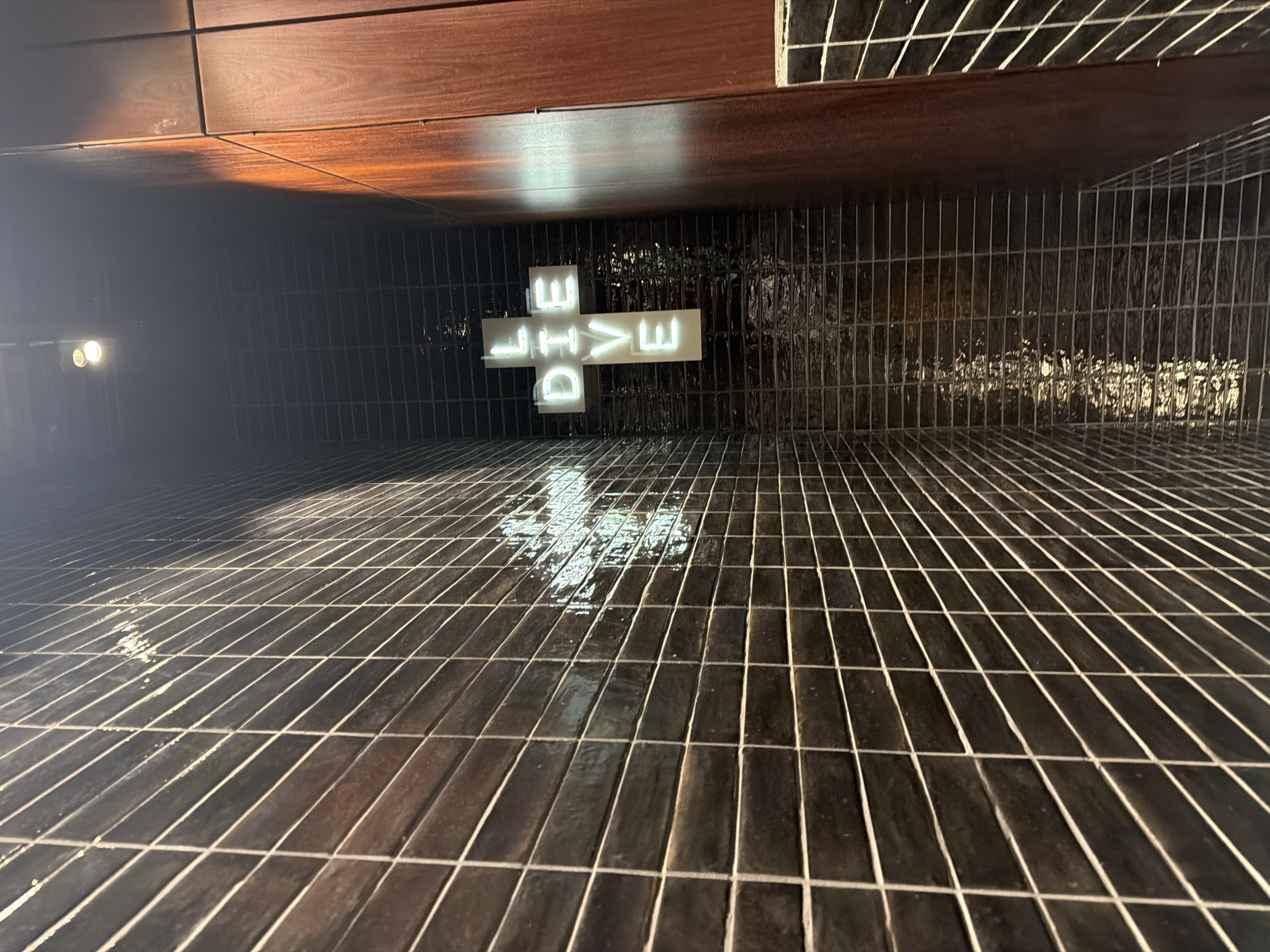 The image depicts the "DIVE" sign in the tiled lobby of the Shangri-La Hotel, located on floors 34-52 of The Shard in London, United Kingdom. The photograph is taken from a low angle, looking upwards. The floor is covered in dark, narrow rectangular tiles with light-colored grout. The tiles reflect the light and the "DIVE" sign above, creating a pattern of diffused reflections. The "DIVE" sign is a neon light installation with the word arranged in a cross shape. Above the sign, there is a dark brown wooden ceiling panel. A pair of lights are visible on the left.
