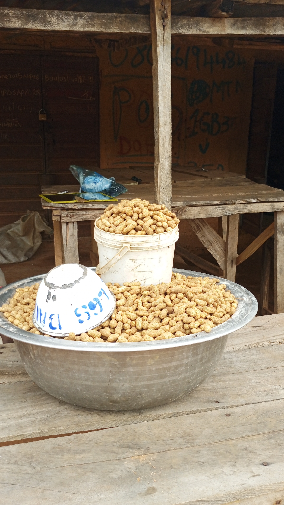 The image shows a close-up view of a large metal bowl overflowing with peanuts. A white bucket, also filled with peanuts, is placed within the bowl, slightly off-center. A white, upturned bowl with blue markings is positioned in front of the bucket and partially submerged in the peanuts. The objects are situated on a weathered wooden surface. In the background, a rustic wooden structure with wooden planks and a wooden post is visible. Some writing in blue and red is present on the wall behind the structure. The overall impression is that of a market stall or an outdoor food vendor in a rural setting. The image is taken from a slightly elevated angle, looking down on the bowl of peanuts. The lighting appears to be natural daylight, with no artificial light sources evident.