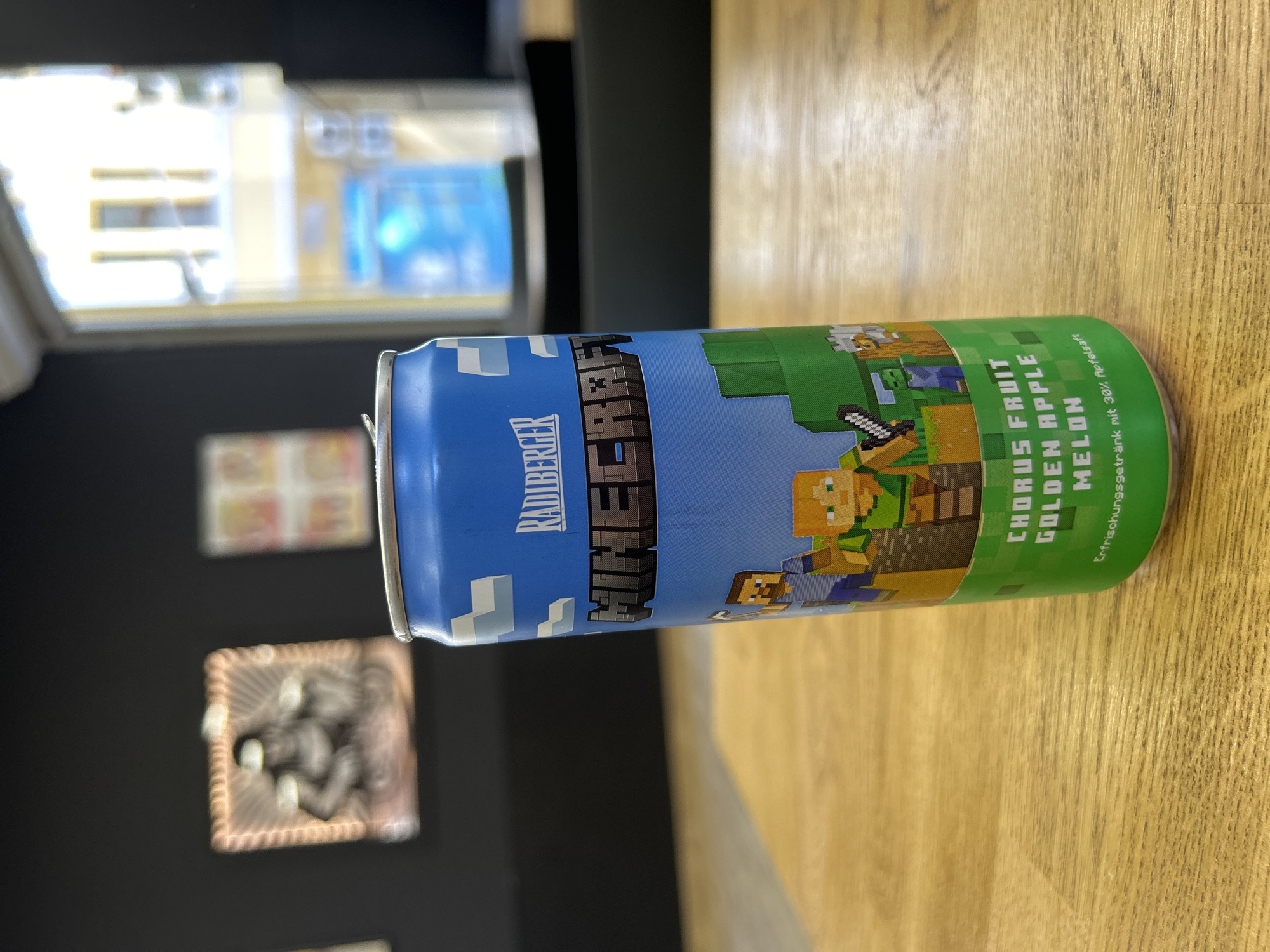 A slightly angled close-up shows a blue and green can of "Radlberger Minecraft" flavored drink standing on a light brown wooden surface. The can has a pixelated Minecraft design featuring characters and landscape elements. The ingredients listed are "Chorus Fruit, Golden Apple, Melon." The background includes a blurred dark gray wall with two framed pictures, and a window with a blurry view of buildings. The location is unknown.
