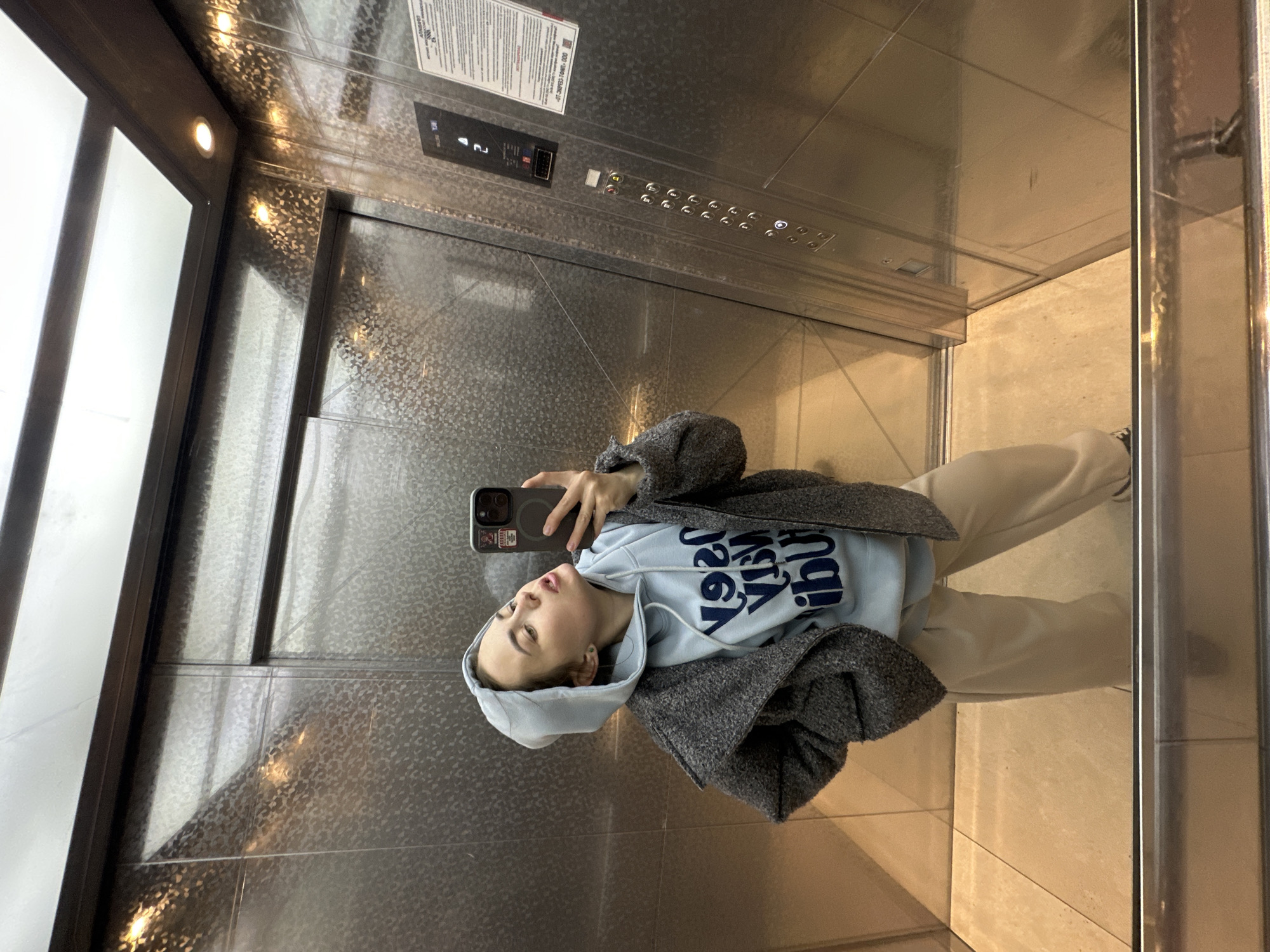 The image shows a woman taking a selfie in an elevator. The elevator has a metal interior, with buttons and a display showing the floor number "4". There is a window on the left side of the elevator. The woman is wearing a grey coat over a light blue hoodie and beige pants. She is holding a smartphone in her right hand, capturing her reflection in the mirrored wall of the elevator. The floor appears to be tiled. The location is indicated by the presence of Russian text near the elevator buttons, and the specific appearance of the surroundings (e.g., floor tiles, door frame) suggests an elevator of Russian construction. Although specific city or locale cannot be pinpointed, Russia can be identified based on observable elements in the image.