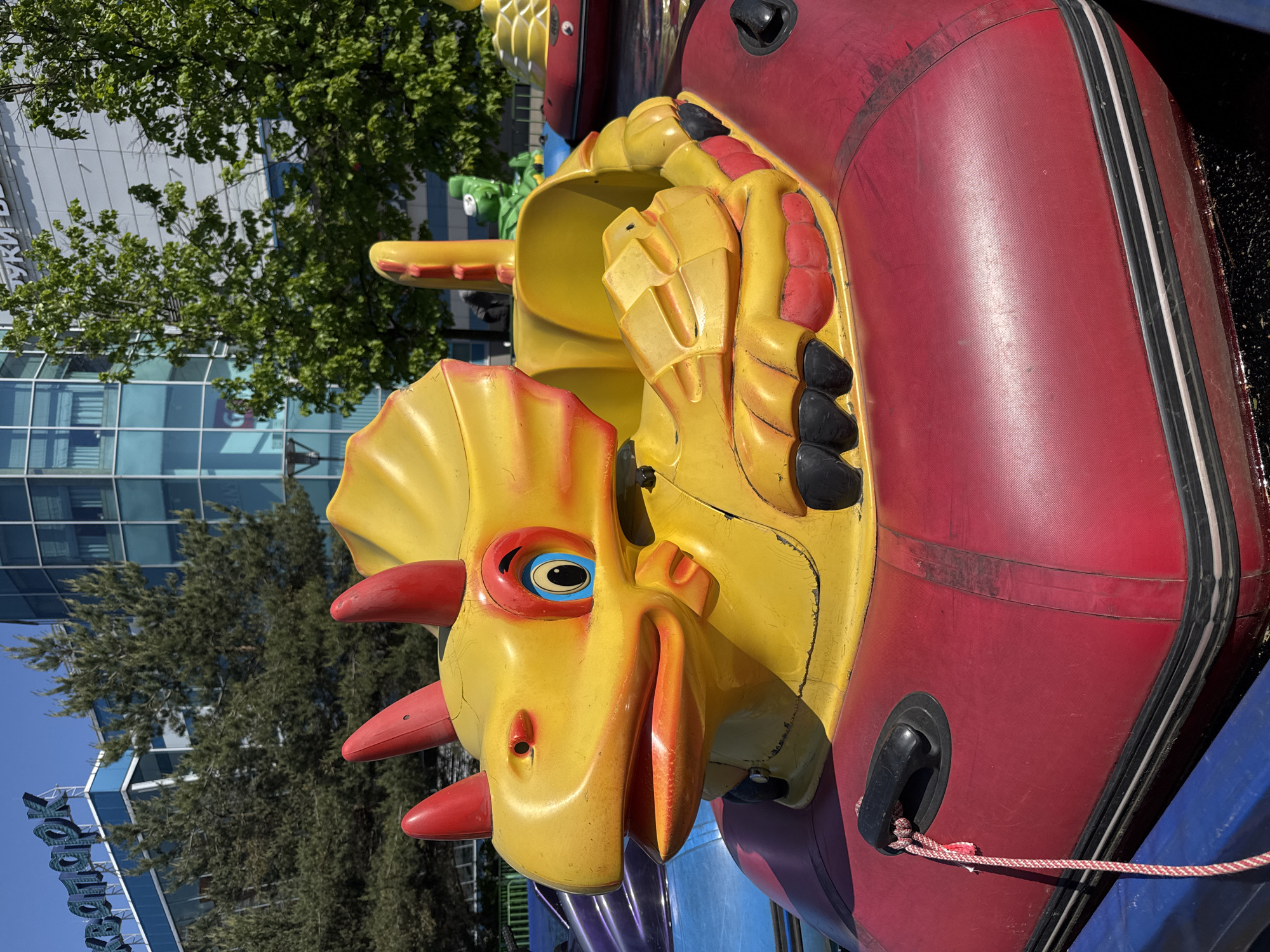An eye-level, medium shot reveals a stationary, Triceratops-themed children's ride at an amusement park in the city of Unknown, Russia. The dinosaur is bright yellow with red horns and facial accents, encased in a red inflatable bumper. The foreground displays a glimpse of a purple and blue base, likely the structure on which the ride is mounted. The background features a modern building with large glass windows and reflective surfaces, partially obscured by trees. The sky is a clear, bright blue, indicating a sunny day. In the lower left corner, signage with the name "Yanauly" is visible.