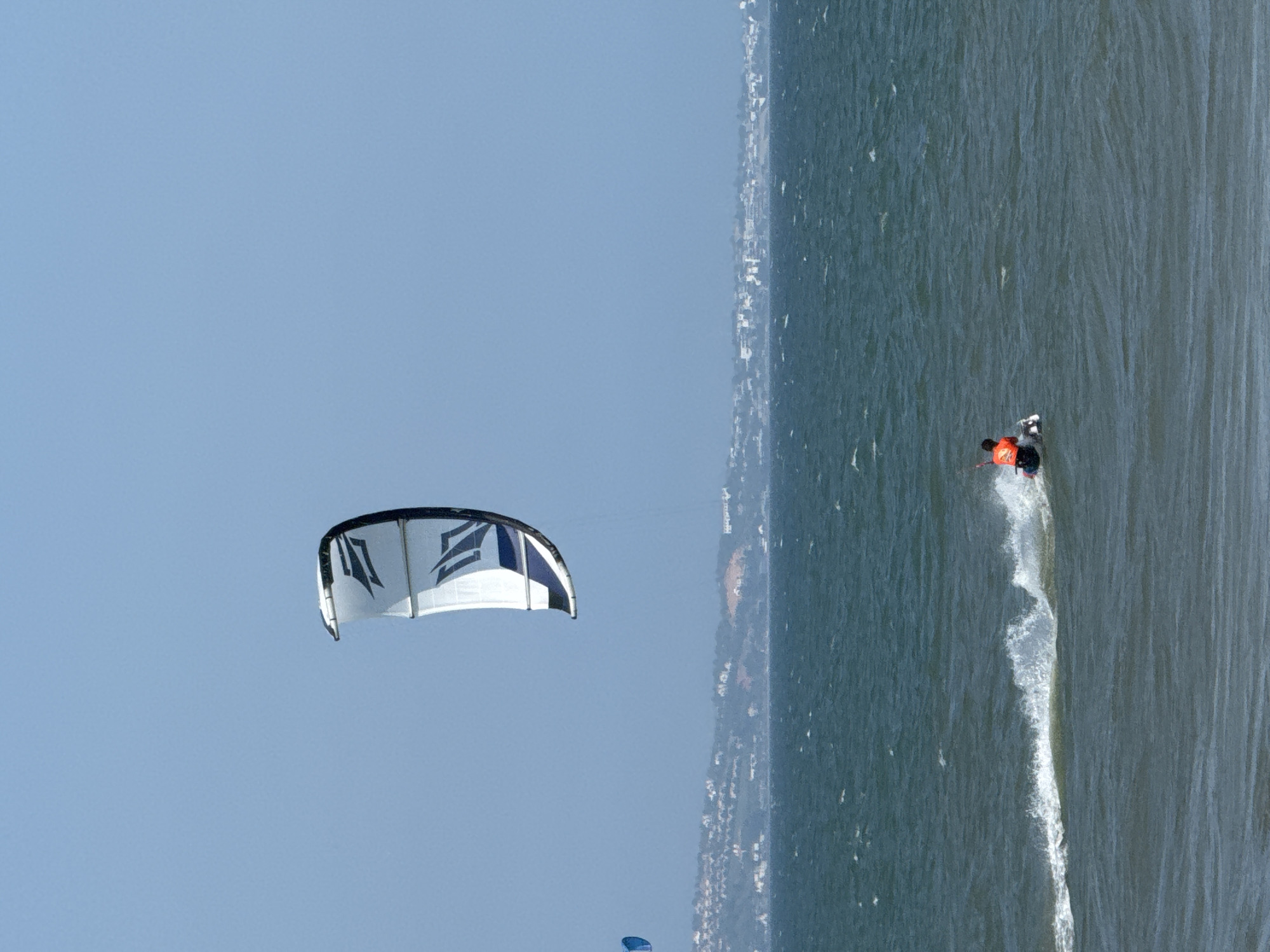 A kitesurfer in an orange vest maneuvers on a choppy, dark blue ocean under a clear, pale blue sky. The kitesurfer, positioned on a white board, is actively engaged in the sport, with a white and blue kite soaring above. In the background, a distant, hazy coastline is visible, dotted with buildings and vegetation, suggestive of a settlement. A smaller blue kite is faintly visible on the left side of the frame, further out in the water. The scene is captured during daylight, likely under sunny conditions, indicated by the bright sky and the white spray kicked up by the kitesurfer. This image depicts a typical watersport activity in a coastal environment, possibly in Phan Thiết, Vietnam, given the visual cues of the coastline and the popularity of kitesurfing in the region.
