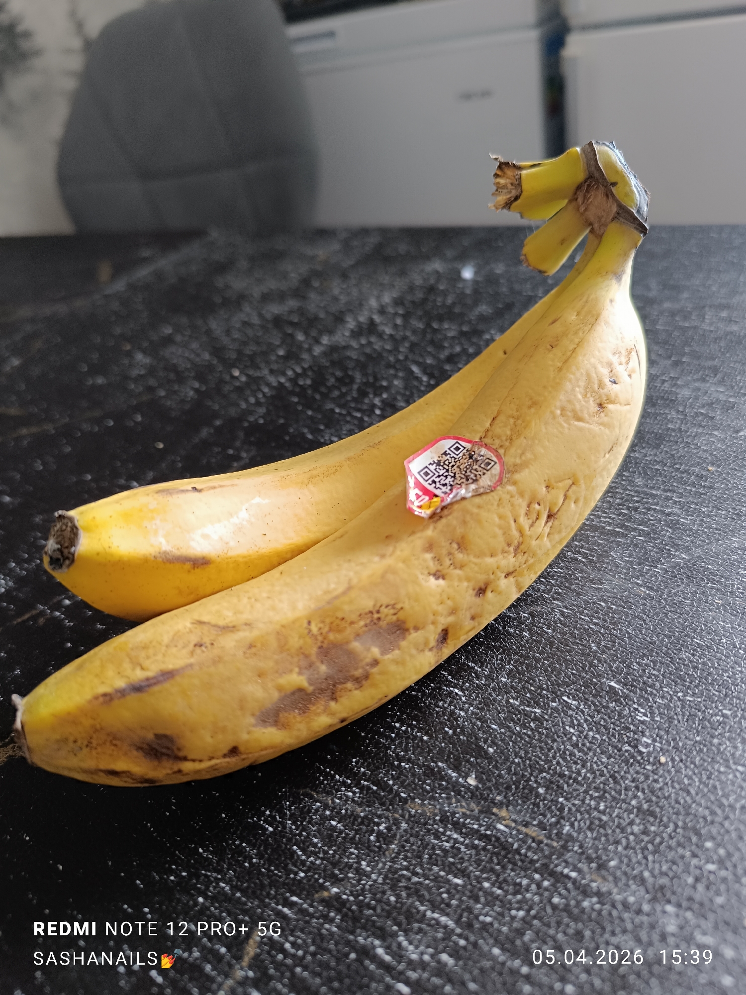 Two yellow bananas are on a black surface. One banana has a sticker with a QR code on it. The location and city details are not provided in the image.