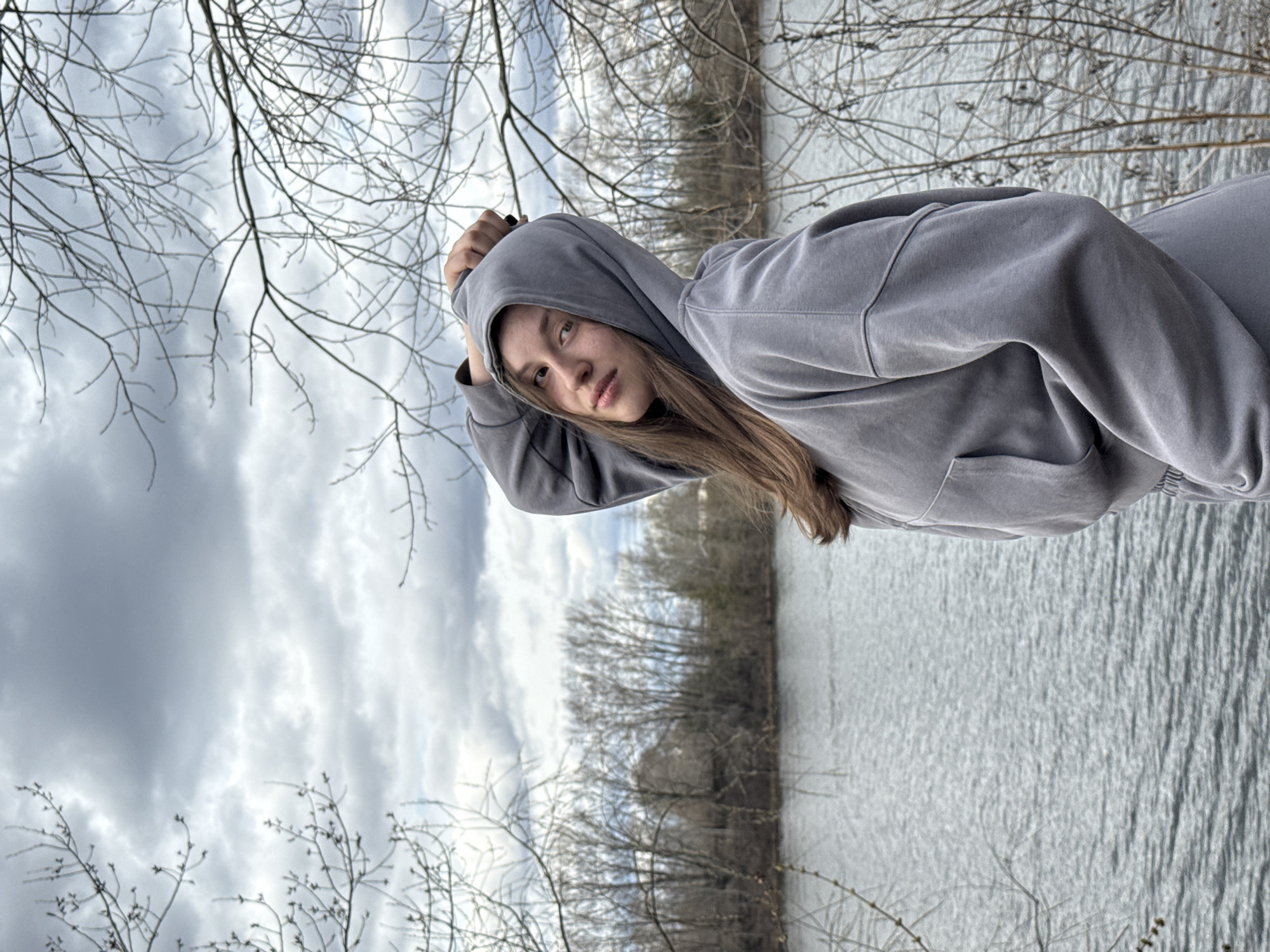 A young woman with light brown hair is wearing a gray hooded sweatshirt and is positioned at an angle in the image, with her head tilted towards the camera. Her right hand is raised near her head, partially covering her hood. The background is a body of water, likely a river or lake, with trees lining the opposite shore. The sky is overcast with gray clouds. Bare branches of trees frame the edges of the image, particularly in the foreground. Based on the description and the features of the landscape, the image is likely taken at or near Chaikovsky, Russia, due to its geographical location near the Kama River and typical climate of the region.