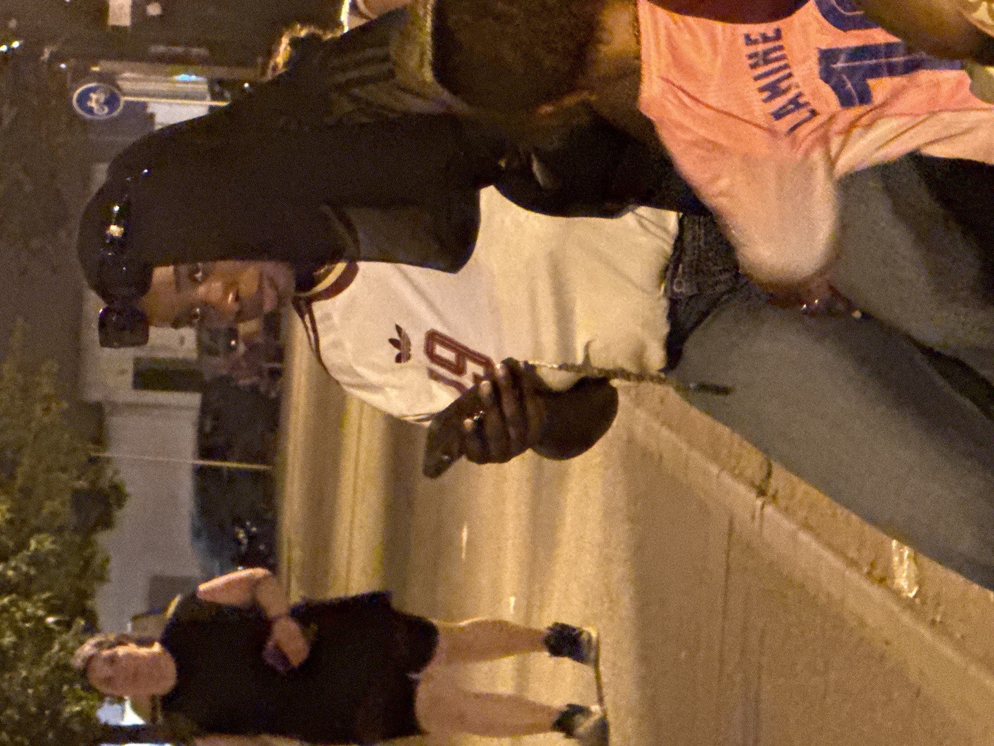 The image depicts a nighttime scene outdoors, possibly on a street in Blanes, Spain, given the context. In the foreground, a person wearing a black niqab and sunglasses is holding a mobile phone in their right hand, appearing to be engaged with it. They are also wearing a white shirt with a maroon trim and a number "19" printed on it, along with blue jeans. To their left, a second person is visible, wearing a black t-shirt and black shorts, with their left arm extended. In the background, another individual is wearing a light pink t-shirt with the text "LAMINE" and the number "1". The environment shows a paved surface, likely a sidewalk, with a street in the background, illuminated by streetlights which cast a warm, yellowish glow. Trees and buildings are partially visible in the blurred background, suggesting an urban setting. A traffic sign with a bicycle symbol is also discernible in the distance.