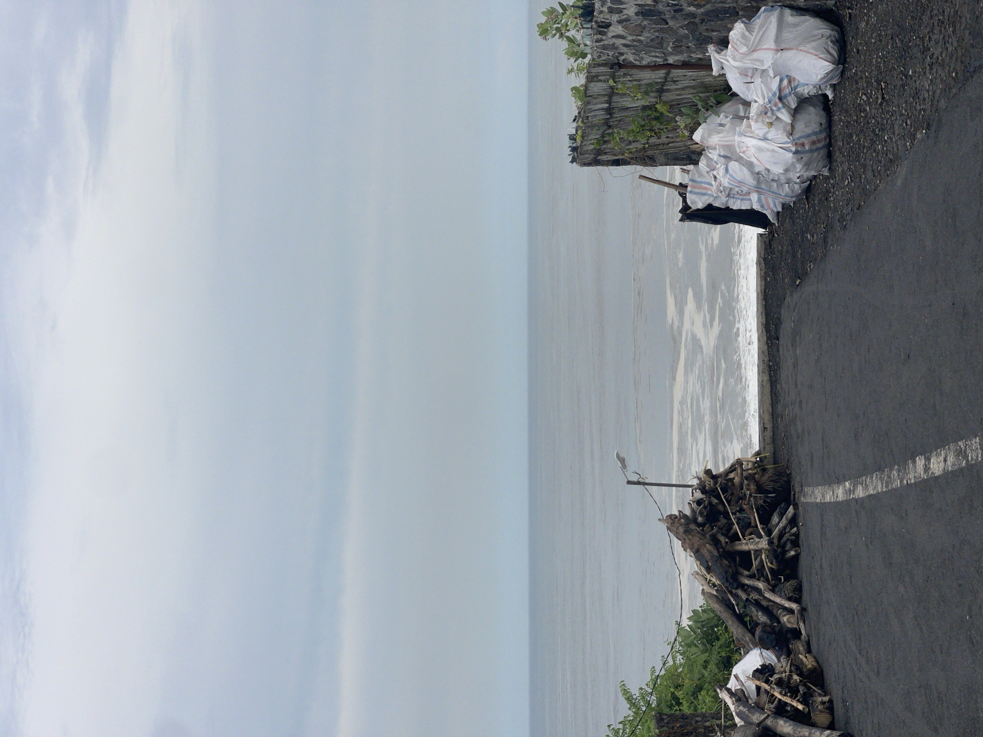 The photograph depicts a coastal scene in Kediri, Indonesia, under an overcast sky. The ocean stretches towards the horizon, meeting the sky in a hazy line. Along the right side of the frame runs an asphalt road with a single white line marking its edge. Adjacent to the road, near the top of the image, there is a pile of large white bags stacked against a low stone wall, with some greenery visible. Further down the road, closer to the bottom of the image, there's a pile of driftwood and debris interspersed with more vegetation. A utility pole extends from the driftwood pile.