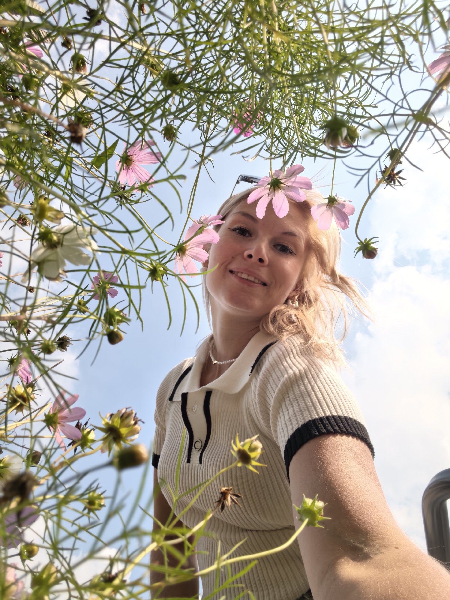 An extreme low-angle medium shot captures a young Caucasian woman with blonde hair, wearing a cream-colored collared shirt with black trim, smiling at the camera. She has a pearl necklace and earrings. Pink cosmos flowers are arranged in her hair and some are in the foreground and background, some in bloom and some as seed heads. The sky is visible in the background with some clouds. The scene appears to be an outdoor flower field or garden. No specific landmarks from Sterlitamak, Russia are identifiable.