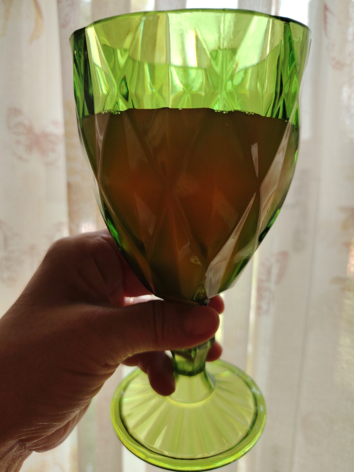 A hand is holding a green glass goblet filled with a dark amber liquid. The goblet has a faceted design, and the liquid appears to have some small bubbles at the surface. The background consists of a sheer, light-colored curtain with a subtle floral pattern, suggesting an indoor setting, possibly near a window. The lighting indicates daylight. The location of Кривец, Russia is not evident from the image content.