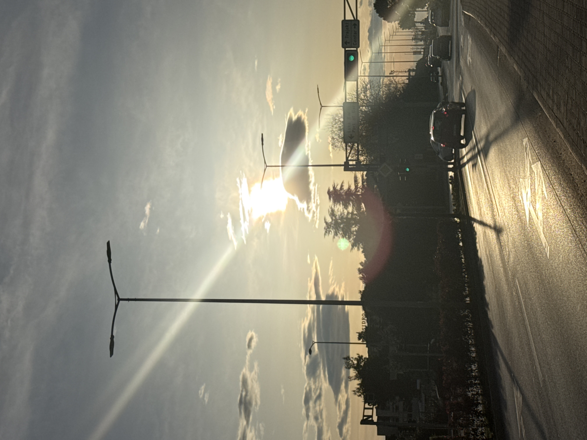 The image shows a road in Bar, Montenegro, with cars driving along it. The sun is setting or rising, creating glare and silhouetting some of the objects in the scene. There are streetlights lining the road. A sign indicating directions to "Trojekt Centar" is visible. The road is marked with arrows indicating the direction of travel. Buildings and trees are visible on the side of the road.