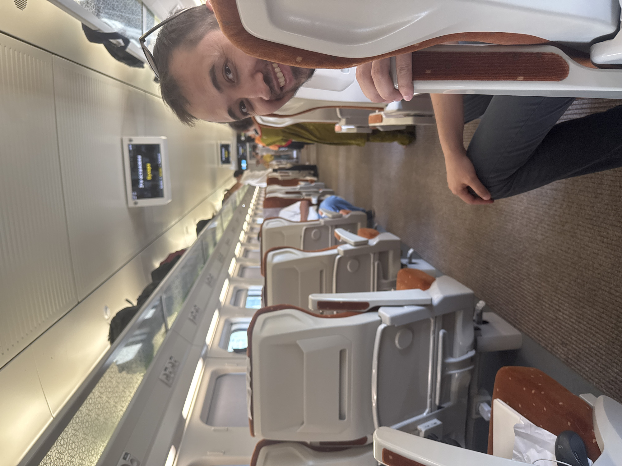 The image shows the interior of a modern train carriage, likely in Uzbekistan. The primary subject is a man with a beard and glasses, who is smiling and looking at the camera. He is sitting in one of the train seats, and the camera appears to be positioned behind him, looking forward down the aisle.
The train carriage has rows of comfortable-looking seats, upholstered in a neutral color with orange accents. The seats are arranged in a configuration that suggests a long-distance journey, with ample legroom and overhead compartments. There are other passengers visible further down the carriage, some appearing to be sleeping or resting. The lighting in the carriage is bright and even, suggesting it's daytime.
On the wall of the carriage, a small screen displays text in Cyrillic script. It appears to be a route map or information display. The overall impression is one of a well-maintained and modern train, likely designed for passenger comfort. The carpeted floor and patterned accent on the wall suggest attention to detail in the carriage's design. The man's relaxed and happy expression conveys a sense of ease and enjoyment of his journey.