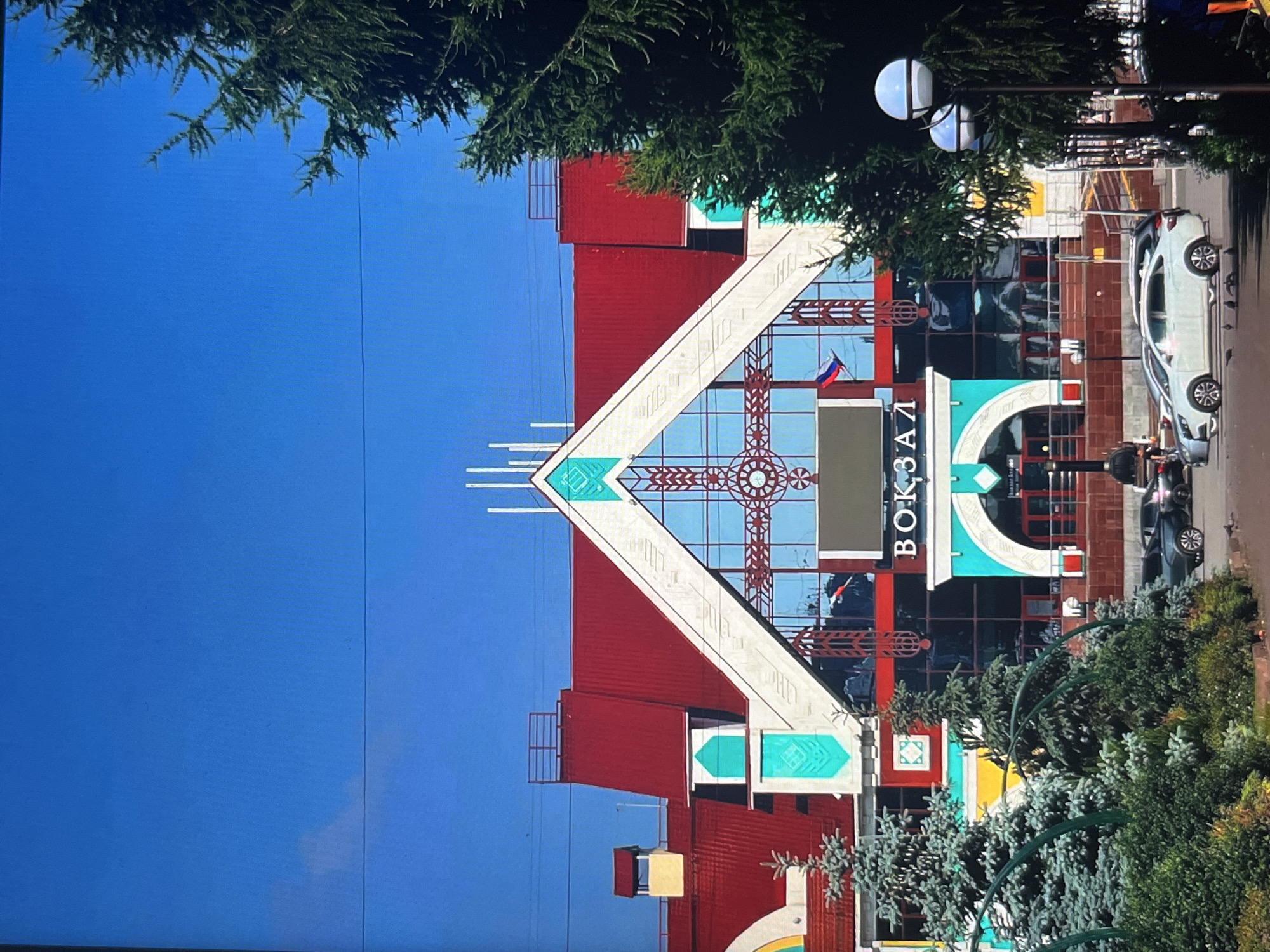 The image shows a modern building with a red roof and large glass windows, featuring bright turquoise accents and a prominent sign that reads "ВОКЗАЛ" (VOKZAL), meaning "station" in Russian. The building is likely a train station in Krasnoyarsk, Russia. The facade is decorated with geometric patterns, and a Russian flag is visible on the building. In the foreground, there are several cars parked, including a white sedan and a dark-colored SUV. Two pine trees are positioned to the right of the building, and smaller evergreen bushes and trees are in front of them. A lamppost with two globes is also visible on the right side of the frame. The sky is clear and blue. The scene appears to be a sunny day in an urban environment.