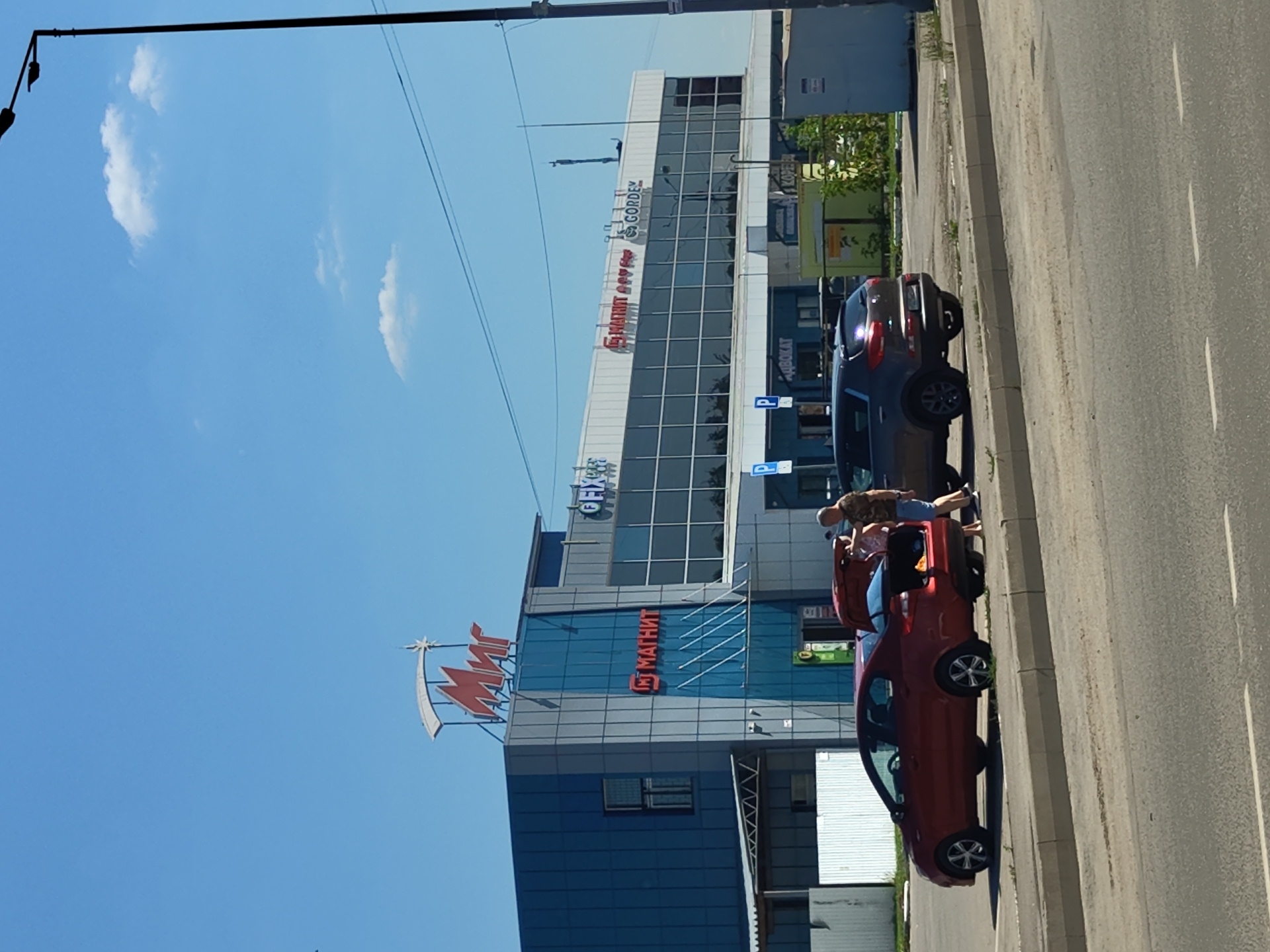 The image shows a sunny day in front of a retail complex in Zhukovsky, Russia. The complex has a large "Мир" (Mir) sign at the top, with the "Магнит" (Magnit) logo visible on the lower part of the building. Several cars are parked in front of the complex. A red sedan has its trunk open, and two people, a man and a woman, are standing near it. The man is wearing a camouflage t-shirt and shorts, while the woman is wearing a white top and shorts. They appear to be engaged in conversation or unloading items from the car. A dark gray SUV is parked next to the red sedan. The environment is urban, with a paved road and sidewalk in the foreground, and the retail complex and surrounding buildings in the background. A tall street lamp is visible on the right side of the image.