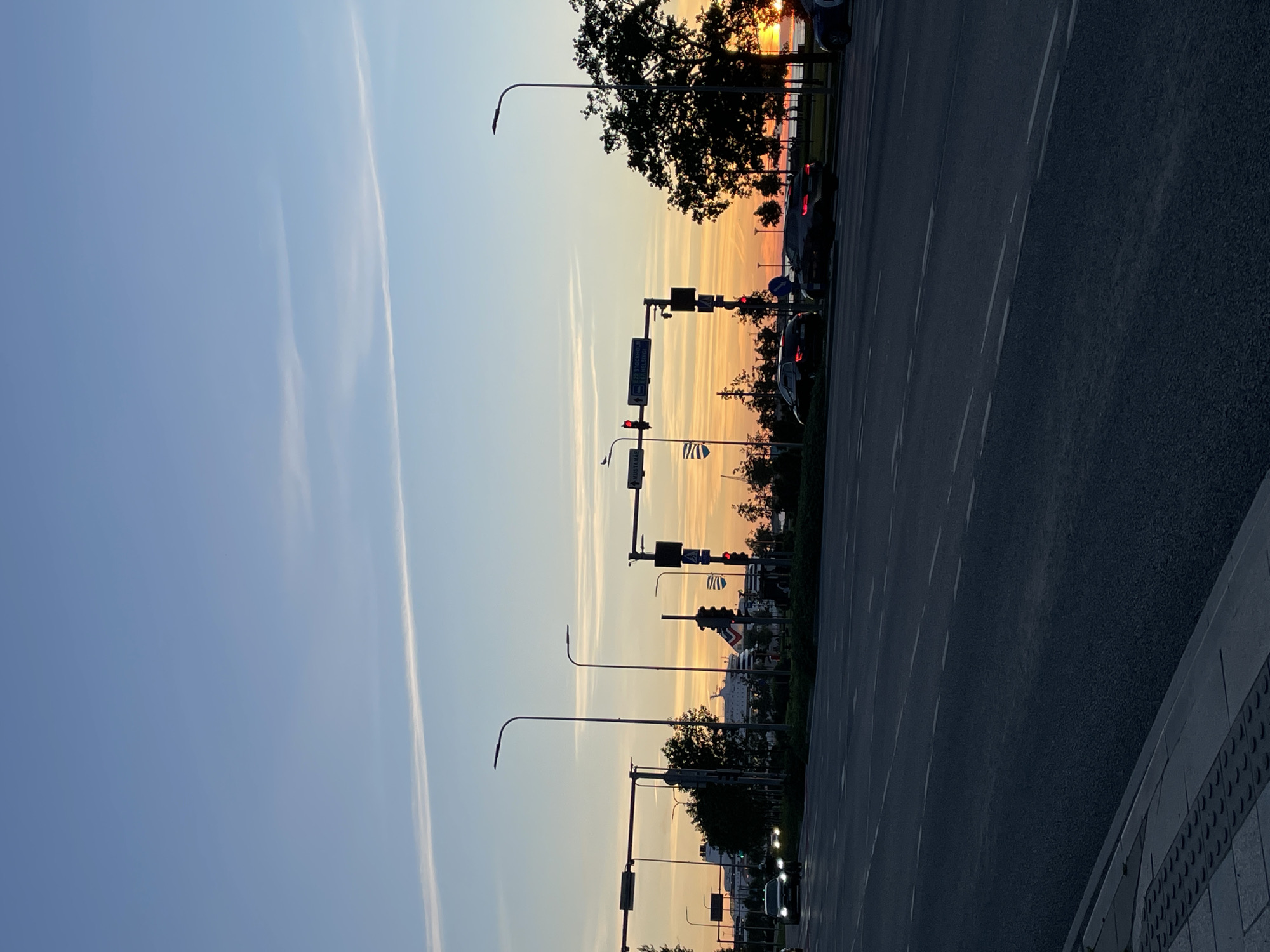 This is a vertical, full-shot, eye-level, outdoor photograph of a sunset on a street in Tallinn, Estonia. The sky is a gradient of pale blue and soft orange, with a thin, white contrail stretching across the upper left quadrant. A street with multiple lanes and white dashed lines runs across the foreground. Several cars, appearing dark and silhouetted, are driving on the street. To the left, tall lampposts with modern streetlights are visible, along with traffic signals. In the midground, there are trees and a large ferry, possibly docked or at sea, with a white hull and red accents. The sunset is casting a warm glow over the scene. The right side of the image features a silhouette of a large tree against the vibrant sunset sky. The overall atmosphere is serene and urban.