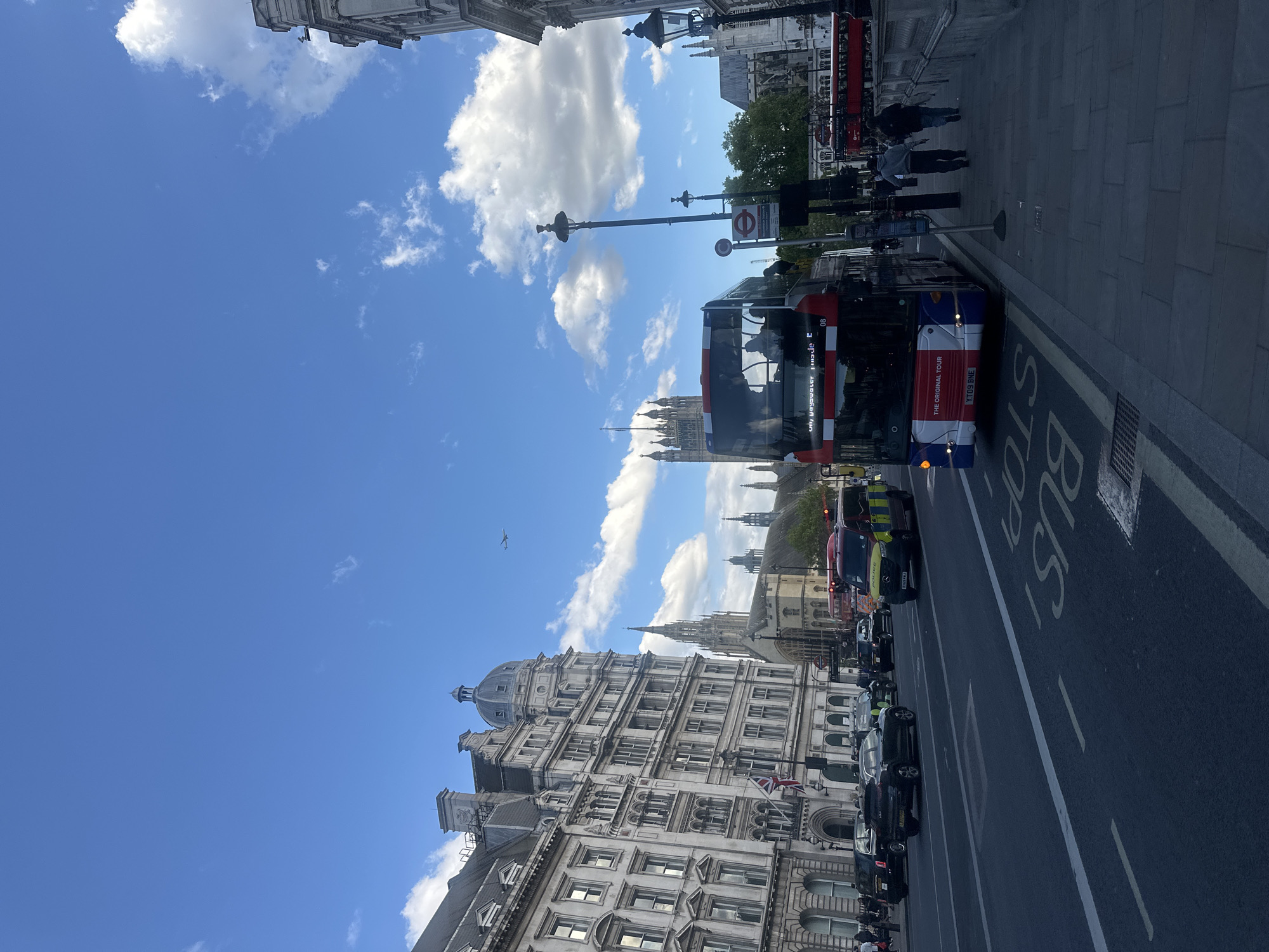 The image captures a street scene in London, United Kingdom, featuring a double-decker bus, traffic, and pedestrians along a city thoroughfare with historic buildings in the backdrop. A red and white double-decker tourist bus is in the bus lane on the right side of the frame. The road has "BUS STOP" painted on it. A line of vehicles, including a police car, is in the lane beside the bus. Pedestrians are walking on the pavement adjacent to the bus lane. Historic buildings with elaborate facades rise on the left side of the frame. The Houses of Parliament are visible further down the street. The sky is blue with puffy white clouds.