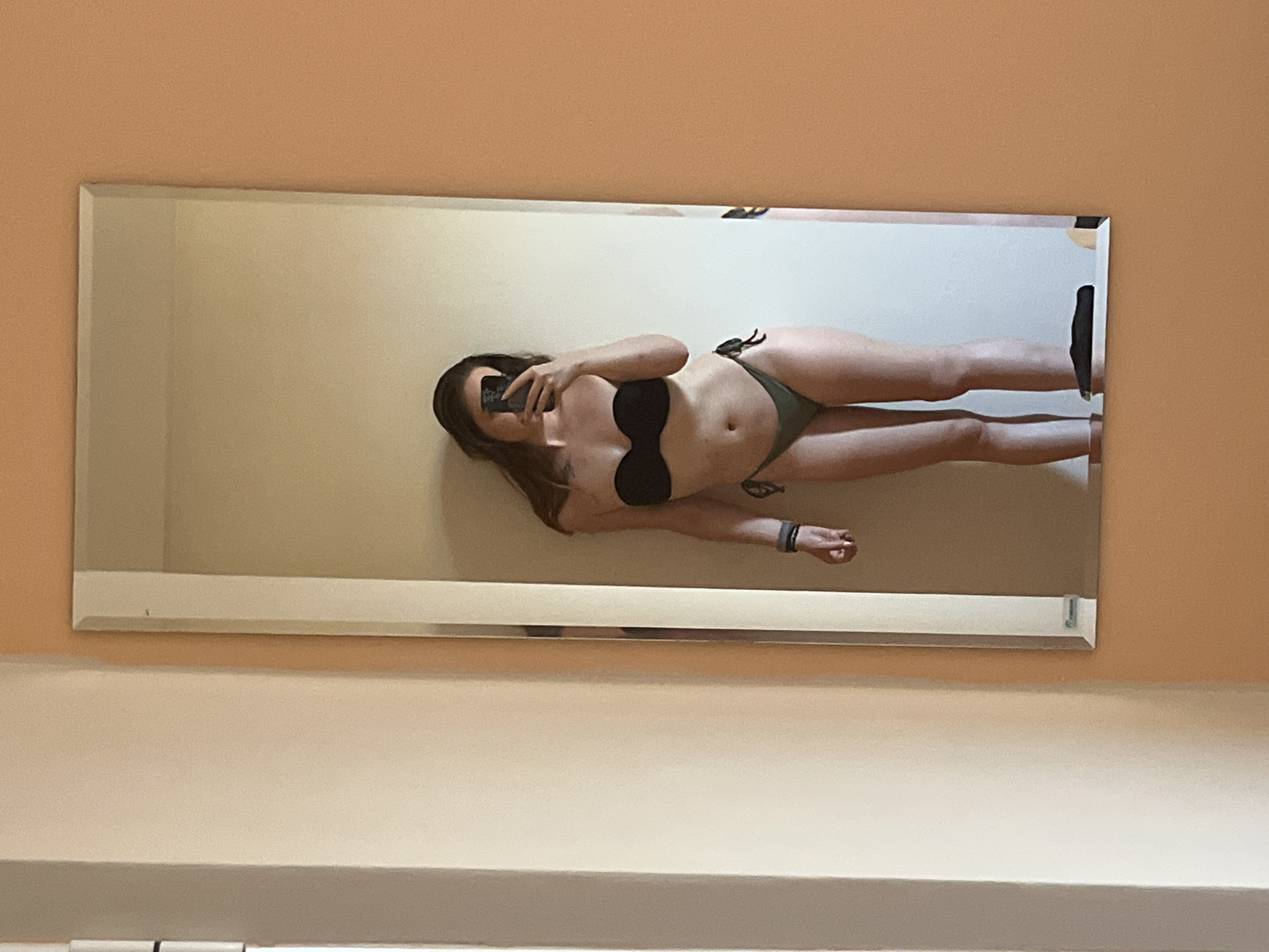 A full-length mirror reflects a person standing in a room. The person is wearing a black strapless bikini top and olive green bikini bottoms. They are holding a smartphone in their right hand, seemingly taking the photo. Their left arm is bent at the elbow, with a dark bracelet or watch on their wrist. The background of the reflection shows a plain wall. The room appears to be a bedroom or a dressing area, with soft, neutral-colored walls and a floor. The lighting is natural, coming from an unseen source, creating subtle shadows. The scene is candid, capturing a moment of self-documentation. There is no discernible information in the image that places it in Lozenets, Bulgaria; the environment appears generic and could be anywhere.