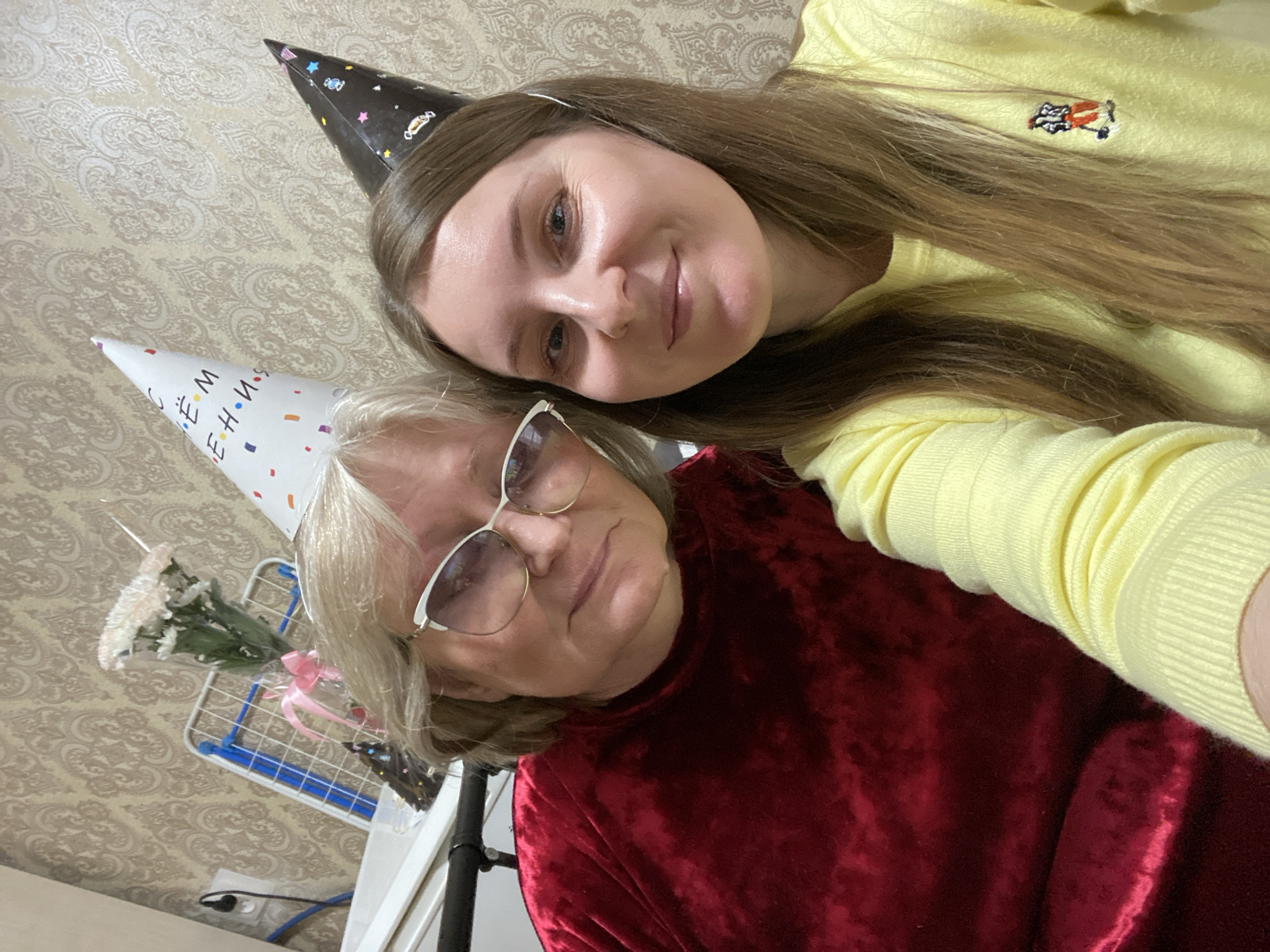 Two females, one older and one younger, are posing for a selfie indoors in Chaikovsky, Russia. Both are wearing birthday hats. The older female has short, white hair and is wearing glasses and a dark red velvet top. The younger female has long, light brown hair and is wearing a yellow sweater. Behind them is patterned wallpaper and a clothes drying rack with flowers on it.