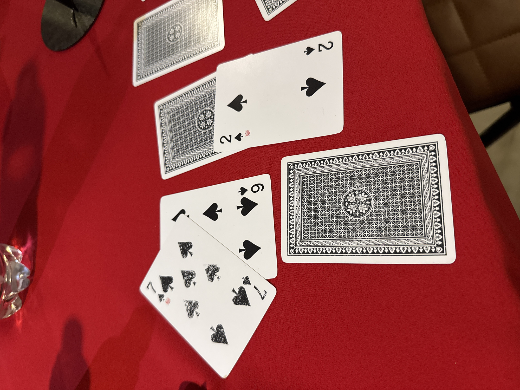 A tabletop view of several playing cards laid out on a red surface. Two cards, a 2 of Spades and another card with its back showing, are positioned in the upper right quadrant. Below them, another card, a 6 of Spades, is partially visible. Further down and to the left, a 7 of Spades is prominently displayed, along with its back. The cards have a classic design, with black spades as the suit symbol. The surface appears to be a felt or cloth material, typical for card games. In the upper left corner, a dark, circular object, possibly a coaster or part of a game accessory, is visible, casting a shadow. A sliver of a glass object, reflecting light, is present in the lower left corner, suggesting a drink or a decorative item. The lighting is warm and direct, creating shadows and highlighting the textures of the cards and surface. The scene is static, implying a paused moment in a card game or a display of cards. There is no visible text on the cards or surroundings other than the standard playing card markings.
