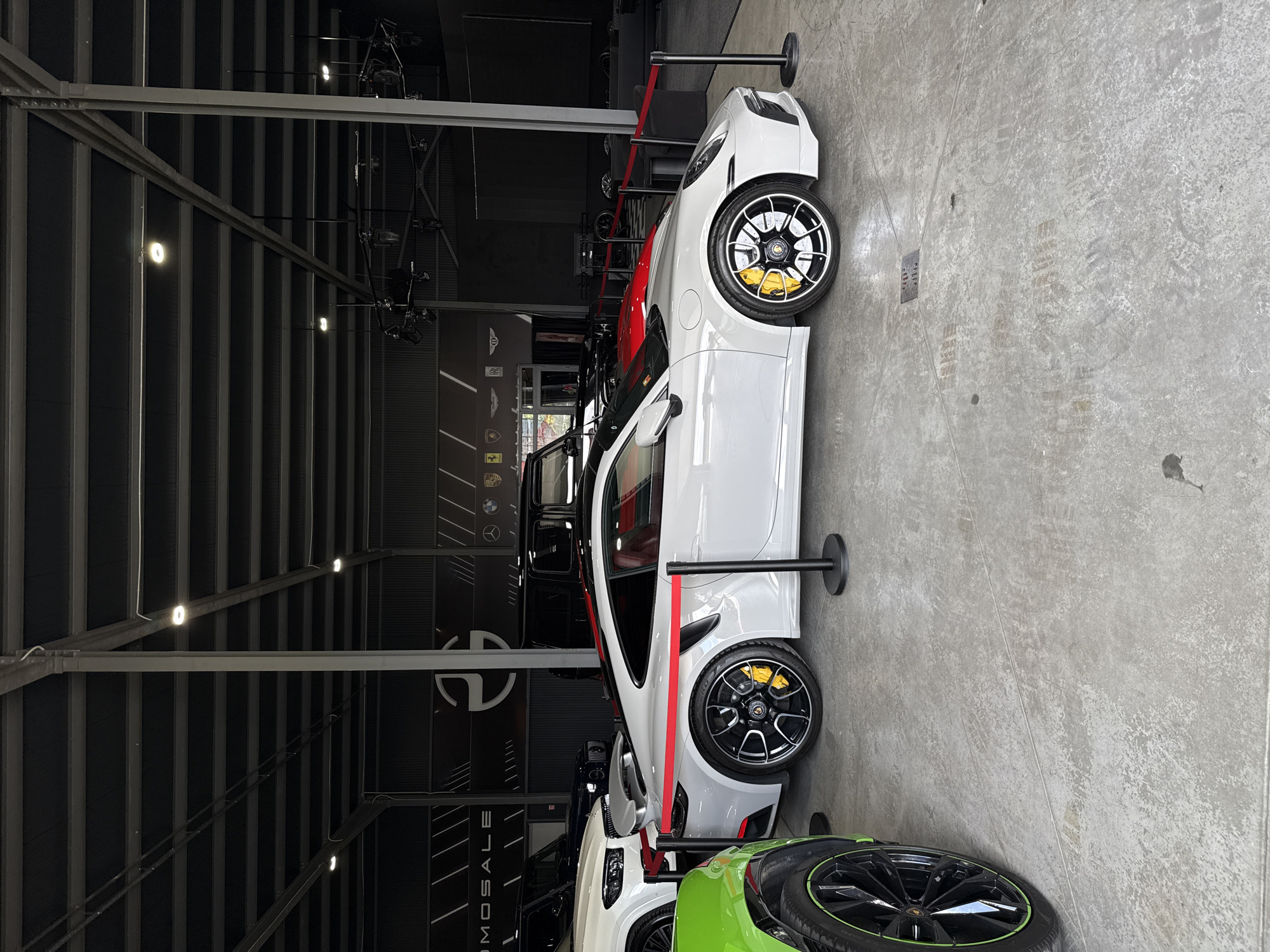 This image displays an indoor showroom or gallery in Plovdiv, Bulgaria, featuring an exclusive display of high-performance sports cars. The primary subject is a sleek white sports car, likely a Mercedes-AMG GT variant, prominently positioned with its left side visible. It features a bold red accent stripe running along its lower body and is equipped with black multi-spoke wheels that highlight bright yellow brake calipers.
Behind the white car, a portion of a red sports car is visible. In the foreground, partially cropped, is a vibrant green car with black wheels featuring a distinctive green rim accent, possibly a Lamborghini. Black stanchions connected by red velvet ropes create a barrier around the vehicles, suggesting an exhibition or premium display.
The setting features an industrial aesthetic with a high, dark ceiling supported by exposed metal beams and trusses, fitted with recessed lights that brightly illuminate the space. The floor is light grey concrete, showing subtle tire marks and general wear.
On a dark wall in the background, several luxury car manufacturer logos are prominently displayed, including Porsche, Mercedes-Benz, BMW, Aston Martin, Ferrari, and Lamborghini, underscoring the high-end nature of the exhibited vehicles. Vertical text reading "AMOSALE" is also visible on this wall, possibly part of a dealership or event name. No people are present, indicating a static exhibition. The overall impression is one of a sophisticated and well-lit automotive showcase.