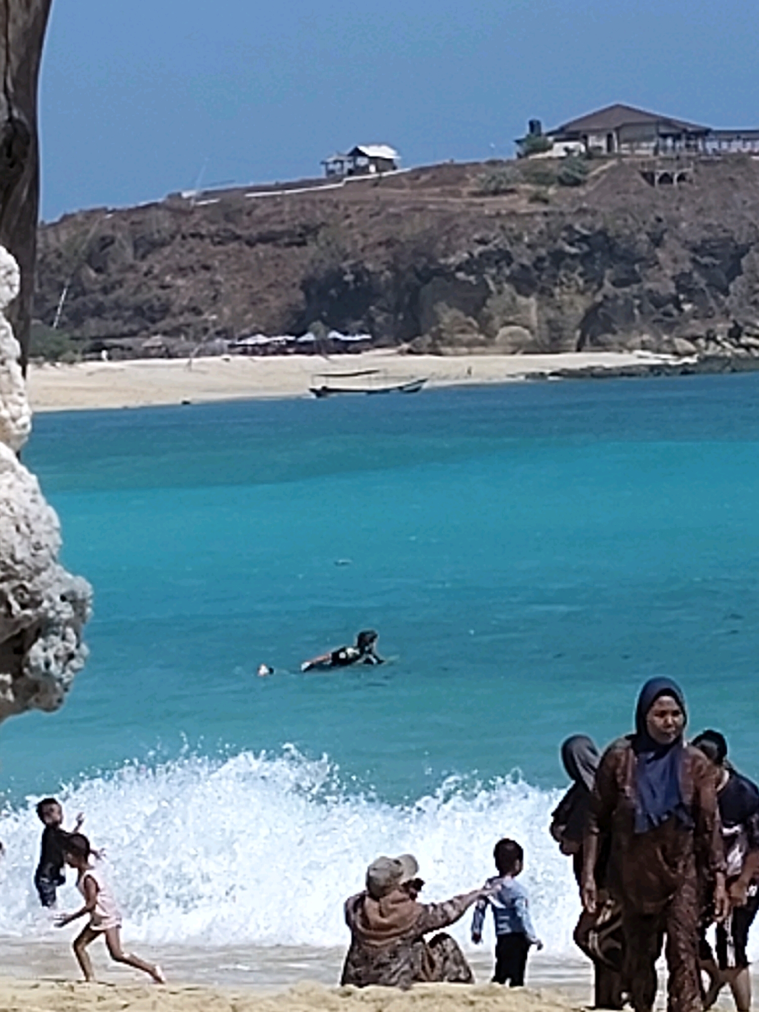 In Kuta, Indonesia, a sunny day at the beach is depicted. The turquoise ocean is active with waves breaking near the shore, where several people are present. In the water, a person is seen lying on a surfboard, while another individual is partially submerged. On the sandy beach, a group of children are playing. One child in a pink dress is running towards the waves, accompanied by another child in a dark top. Further back, a woman in a headscarf and patterned brown clothing is walking, with other people behind her. A person wearing a hat and seated on the sand is reaching out towards a child in a blue shirt. The background features a dry, hilly landscape with buildings and vegetation on top. The overall scene captures a vibrant beach day with various activities and individuals enjoying the coastal environment.