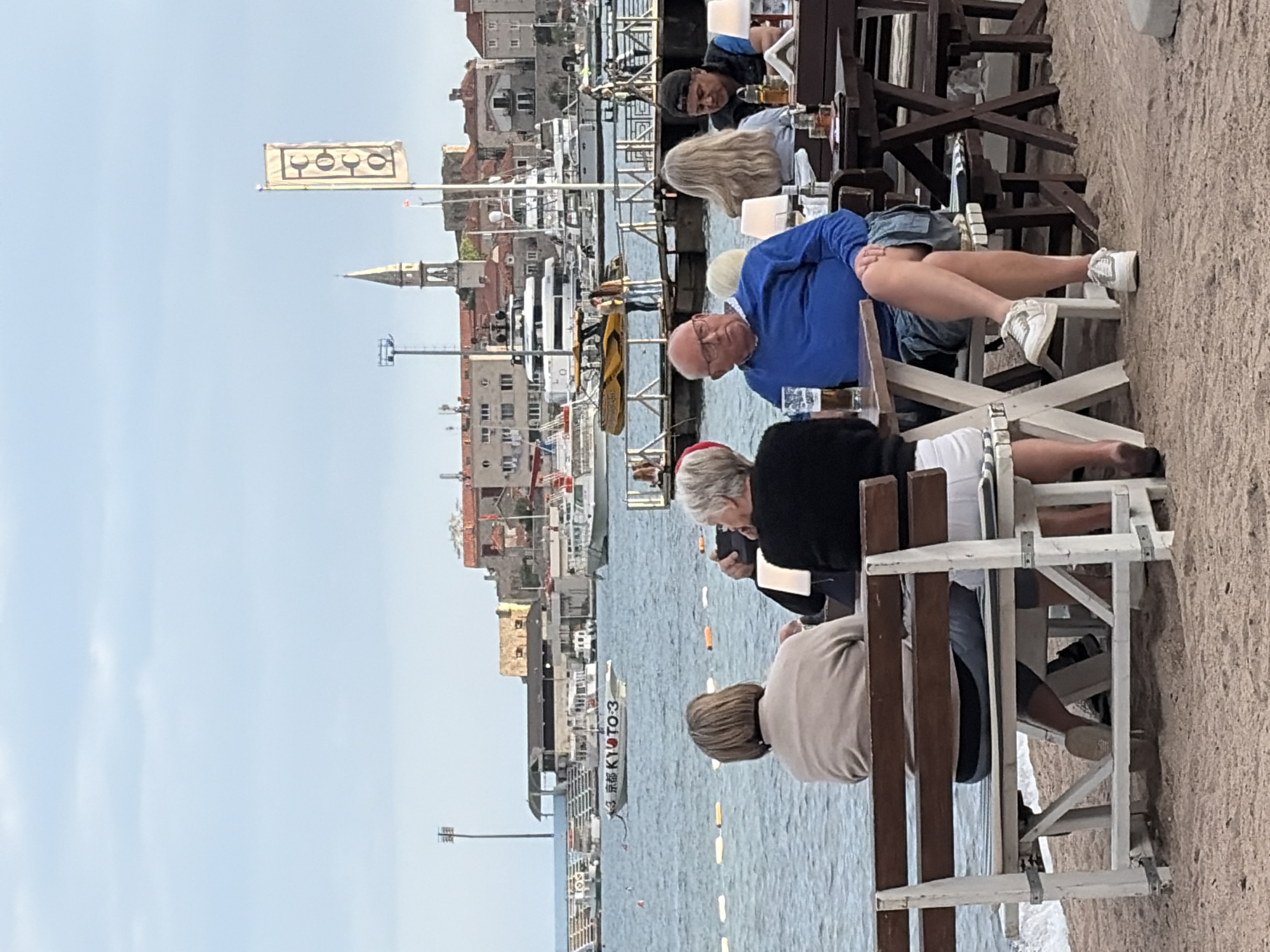 Captured on a sunny day, the image shows a waterside scene likely in Budva, Montenegro, due to the architectural style and beachside setting. A group of people is visible, seated at tables or benches, enjoying a meal or drinks. The group includes men and women of varied ages; one man is wearing a blue shirt and glasses. One woman is wearing a red head covering. They are arranged in tiers, possibly at a restaurant with tiered seating next to the water. In the background, the city of Budva is visible, featuring multiple buildings with orange tiled roofs, a church spire, and a flag with a design of three drinking glasses. Several boats are docked in the marina. The foreground shows a sandy beach.