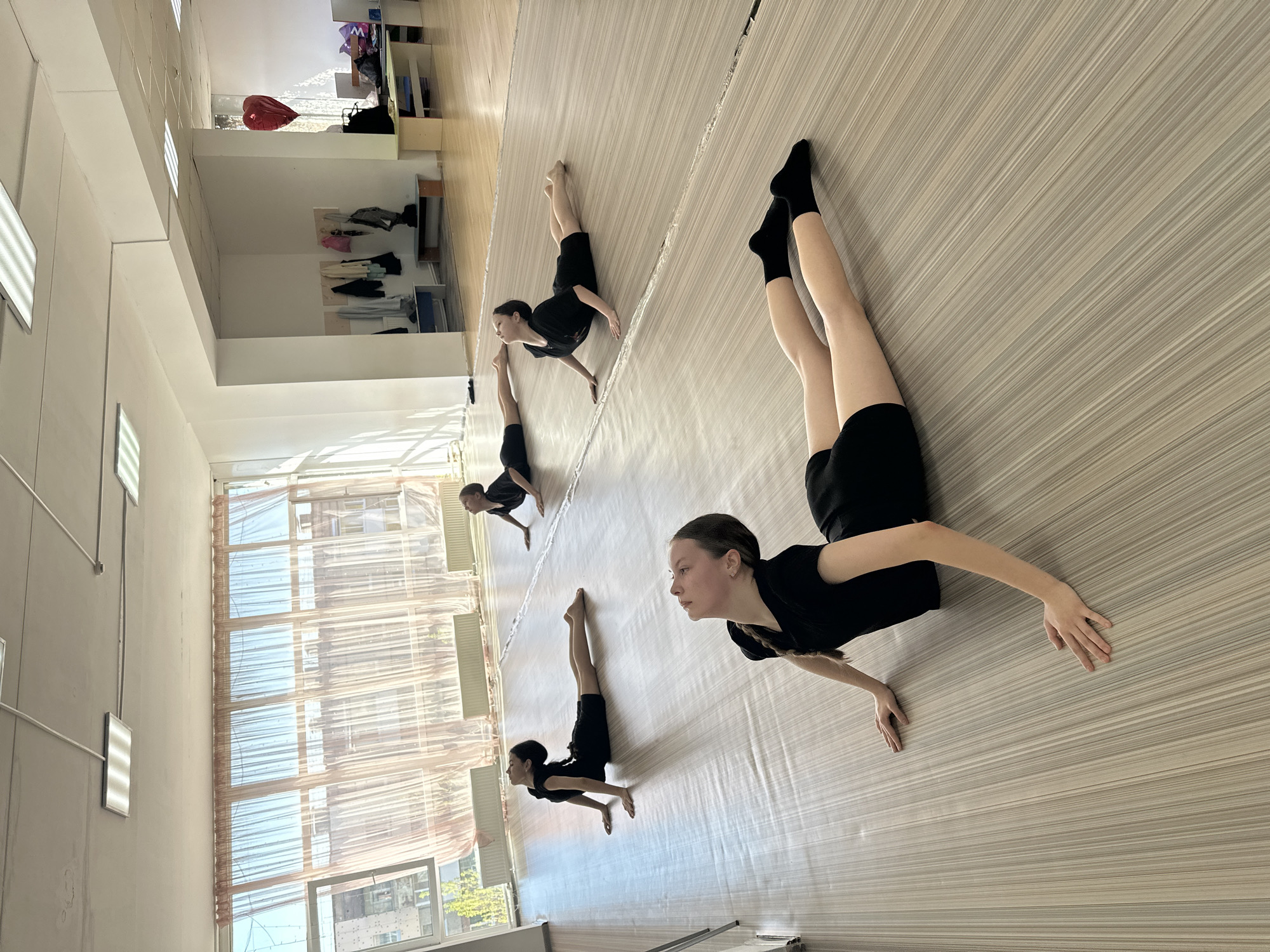 The image shows four young women performing a floor exercise in a dance studio. The studio has a light-colored wooden floor and large windows covered with sheer curtains. In the background, there are shelves with clothes and bags. The women are wearing black shorts and tops, with one wearing black socks. They are positioned in a line, performing a similar movement with their legs raised and arms extended. The location cannot be verified as Votkinsk, Russia.