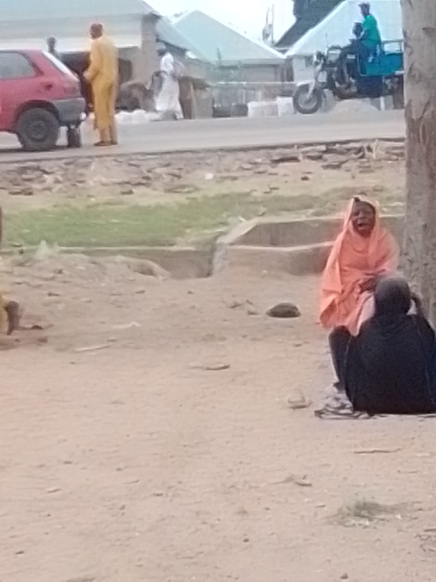 The image depicts an outdoor scene, possibly on a street or dirt road, in Pantame, Nigeria. In the foreground, two individuals are visible. One person, wearing a black headscarf and covering, is seated. Facing them is another individual wearing a bright orange hijab and dress, also seated. The background reveals a red car parked on the side of a road. Further in the background, several people in traditional attire are walking. A blue and green motorized tricycle, commonly known as an 'okada' or 'keke napep', is also seen on the road, with two occupants. The environment appears to be dusty, with some vegetation and concrete structures visible. The overall impression is a snapshot of daily life in a Nigerian urban or semi-urban setting.
