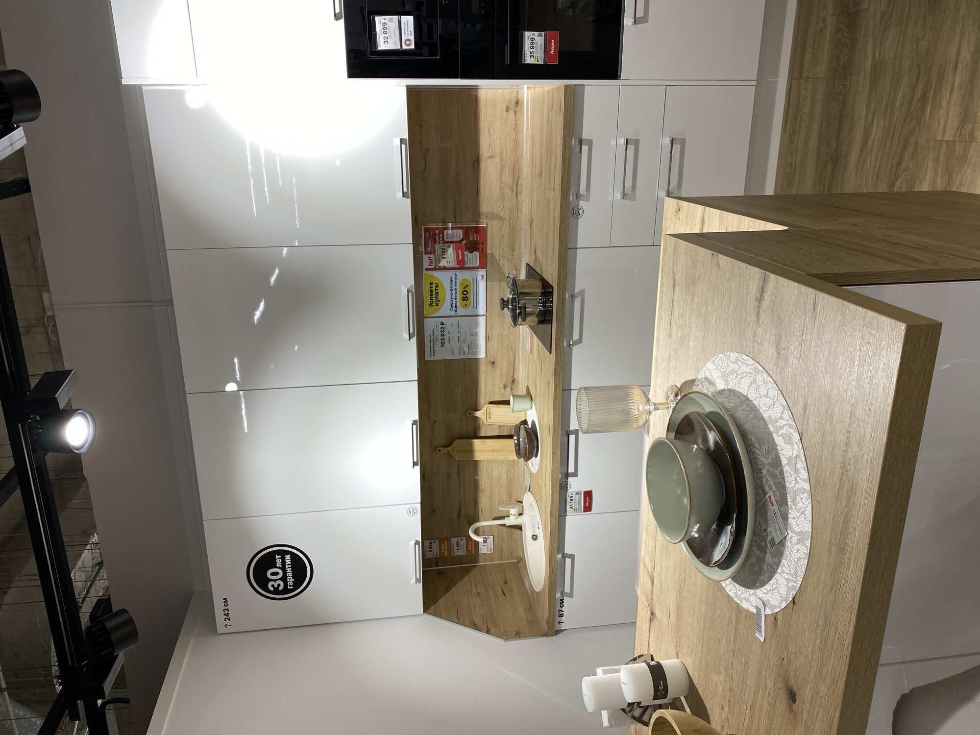 The image shows a kitchen display in a store, possibly in Murino, Russia, given the Cyrillic text on the price tags and warranty information. The kitchen features white cabinets, a faux-wood countertop, and a black cooktop/oven unit. There's a sink and faucet on the countertop, along with some kitchenware and a cooking pot. The countertop also features dishes set on a patterned placemat. There are price tags visible, including details about discounts and promotions. The display is well-lit, likely using spotlighting, with track lighting visible in the upper-left corner of the image. The text suggests a 30-year warranty on the kitchen unit.
