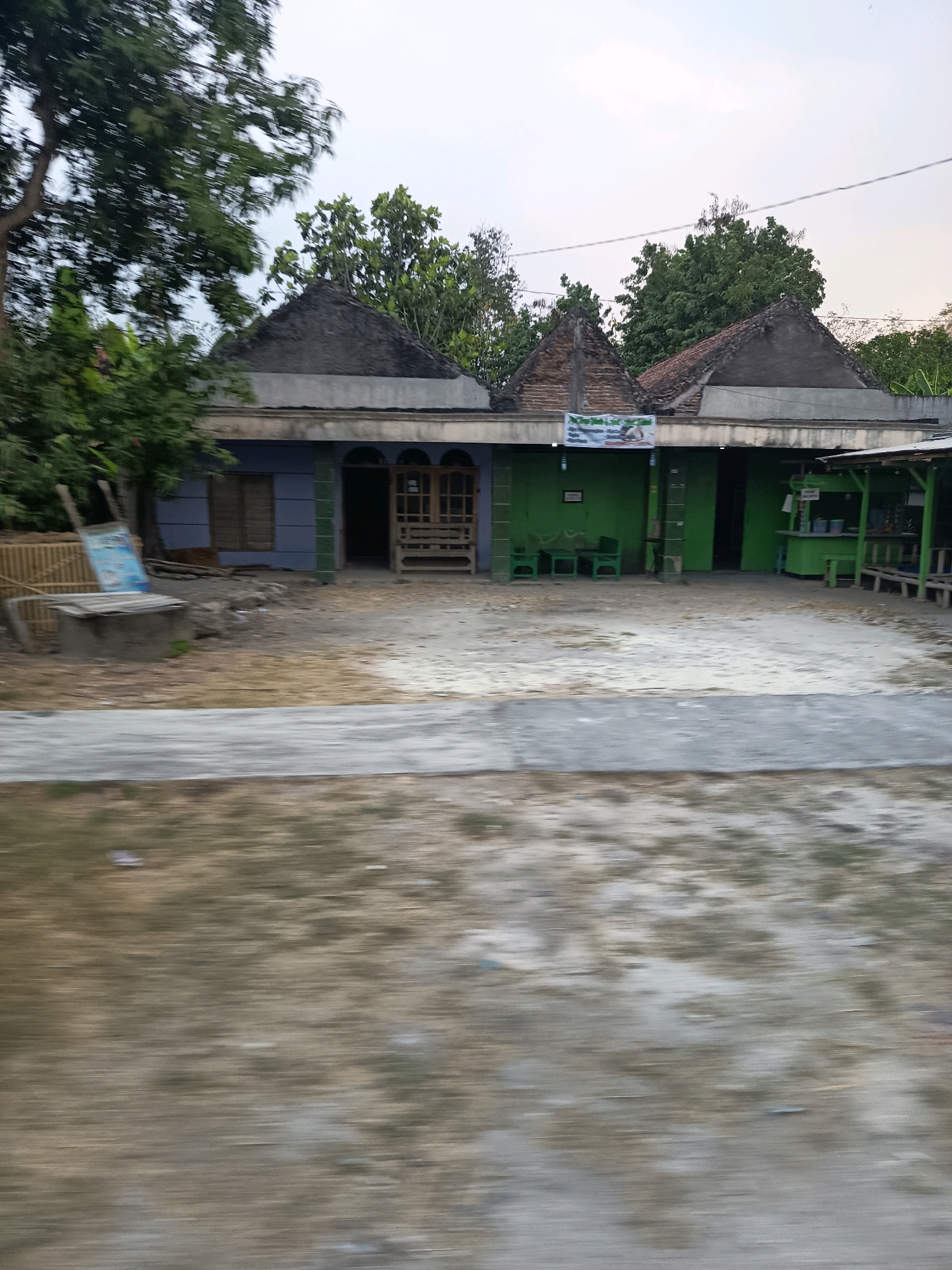 The image shows a roadside scene in Jetaksari, Indonesia, characterized by a blur of motion. In the foreground, a blurred effect suggests movement, with indistinct shapes and colors creating a sense of speed.
Behind the motion blur, a building complex is visible. The structure features multiple rooflines, some with dark, conical roofs and others with more traditional pitched roofs covered in reddish-brown tiles. The walls of the building are painted in various colors, including a pale blue and vibrant green. Several doorways and windows are present, some of which are partially obscured by wooden structures.
In front of the building, a concrete or paved area is visible. There are some green chairs and tables set up outside, suggesting a seating area for a business. A small stall or shop with a green awning is also present, with what appear to be shelves or display areas visible.
To the left of the building, a wooden fence and a large tree are present. There's also a concrete structure that might be a well or a base for something. A sign, likely for a business, is leaning against the fence.
The environment appears to be somewhat rural or suburban, with trees and vegetation surrounding the building. The sky in the background is overcast, suggesting it might be late afternoon or early morning. The overall impression is of a functional, perhaps commercial, establishment on a thoroughfare in Jetaksari.