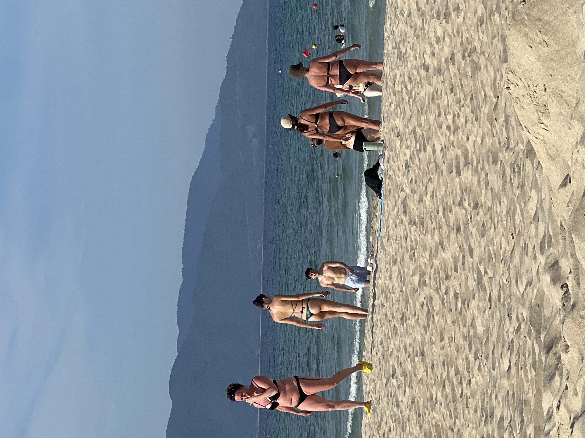 The image captures a sunny day at a beach in Đà Nẵng, Vietnam, with the backdrop of misty, layered mountains. The foreground is dominated by undulating sand dunes. In the mid-ground, several people are visible near the water's edge.
On the left, a woman in a black and white bikini and bright yellow Crocs walks along the sandy shore, her hand near her face. To her right, a woman in a green bikini stands facing away from the camera, looking out at the sea. Next to her, a man in blue swim trunks stands in the shallow water.
Further out in the sea, several other individuals are visible, some swimming or wading. The water is a deep blue, with gentle waves breaking on the shore. Red and yellow buoys are scattered in the water.
To the right of the central figures, a group of women in bikinis are standing closer to the water. One wears a beige hat and a black bikini, another a dark hat and black bikini. Another figure, partially obscured, is also visible. In the water behind them, more people can be seen. The overall scene depicts a leisurely day at the beach with moderate activity.