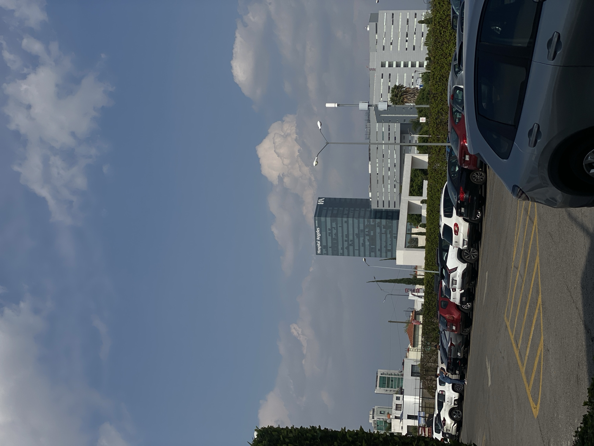The image depicts an outdoor urban or suburban scene on a clear, sunny day. The sky, a dominant feature, is a bright blue with scattered white and light grey clouds.
In the foreground, a parking area or street is visible, marked with yellow lines on asphalt. A row of cars is parked along this area. The closest car to the viewer, on the right side, is a silver-grey hatchback. Further down the row, various other cars are parked, including red, black, and white vehicles. A person is partially visible near a white car, possibly getting in or out.
The midground and background feature several modern, multi-story buildings. A prominent, dark-colored skyscraper stands tall, with its facade displaying reflective windows and the name "Hospital Ángeles" clearly visible in white text. To the right and behind this building, a complex of lighter-colored, contemporary buildings with many windows can be seen. Green hedges and various trees, including palm trees and conifers, are interspersed throughout the scene, bordering the buildings and parking areas.
The scene suggests an environment around a hospital or a commercial complex, characterized by modern architecture and ample parking under a bright sky. No specific activity beyond cars being parked and one person interacting with a car is evident.