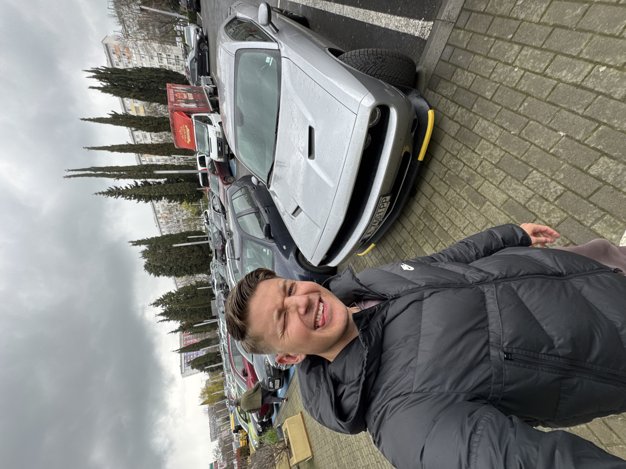 A man is standing outdoors on a brick-paved surface, seemingly in a parking lot in Burgas, Bulgaria, based on the license plate A 7601 PP visible on the car. He is wearing a dark, puffy jacket and is making a playful face, squinting his eyes and sticking out his tongue. Behind him, several cars are parked in a row, including a silver Dodge Challenger with yellow bumper guards. Trees line the background alongside buildings. The sky is overcast.